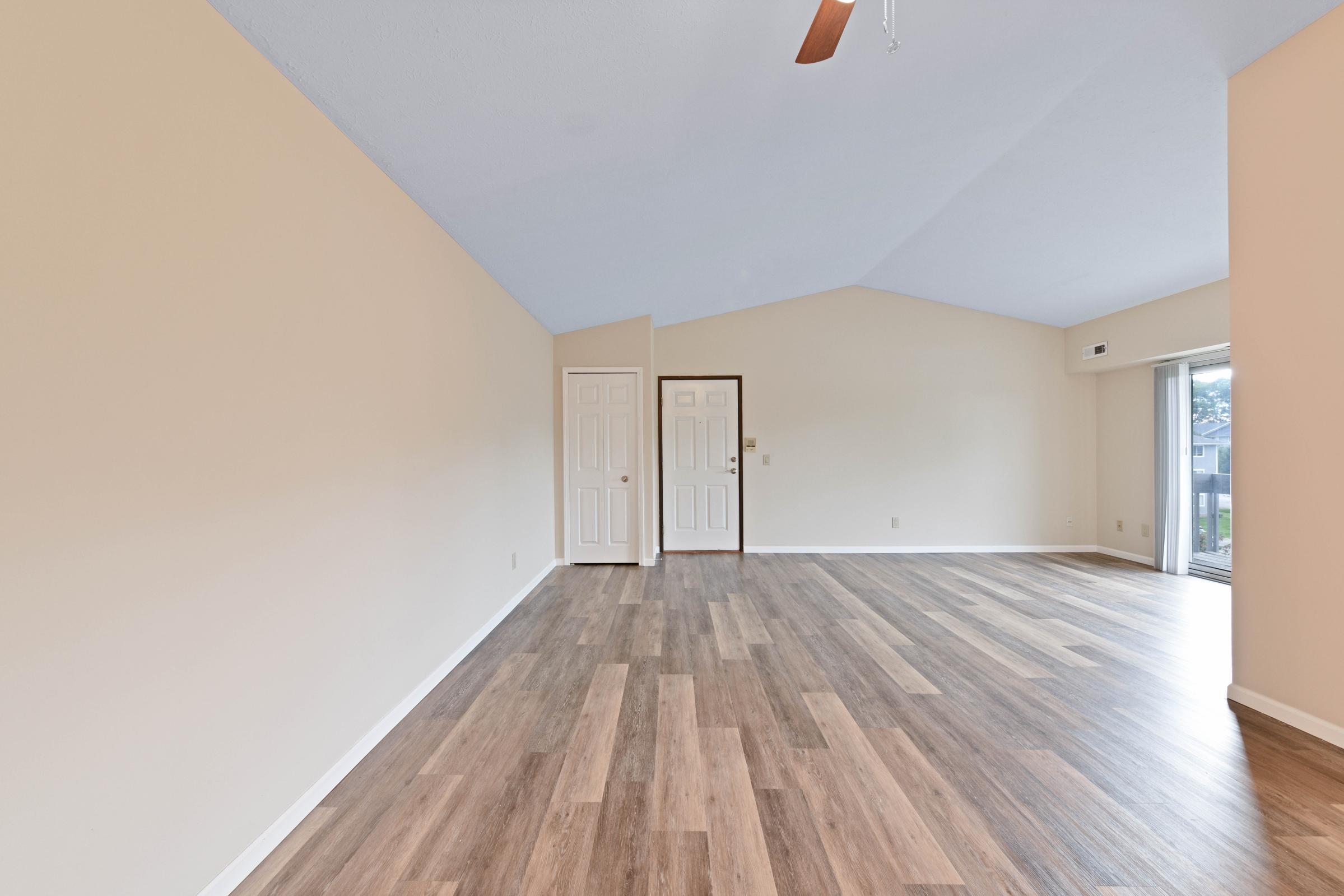 A spacious room with high ceilings and light-colored walls. The floor features wood-like laminate, and there are two closed doors on one side, leading to another area. A large sliding glass door on the right opens to an outdoor space. Natural light fills the room, creating a bright and inviting atmosphere.