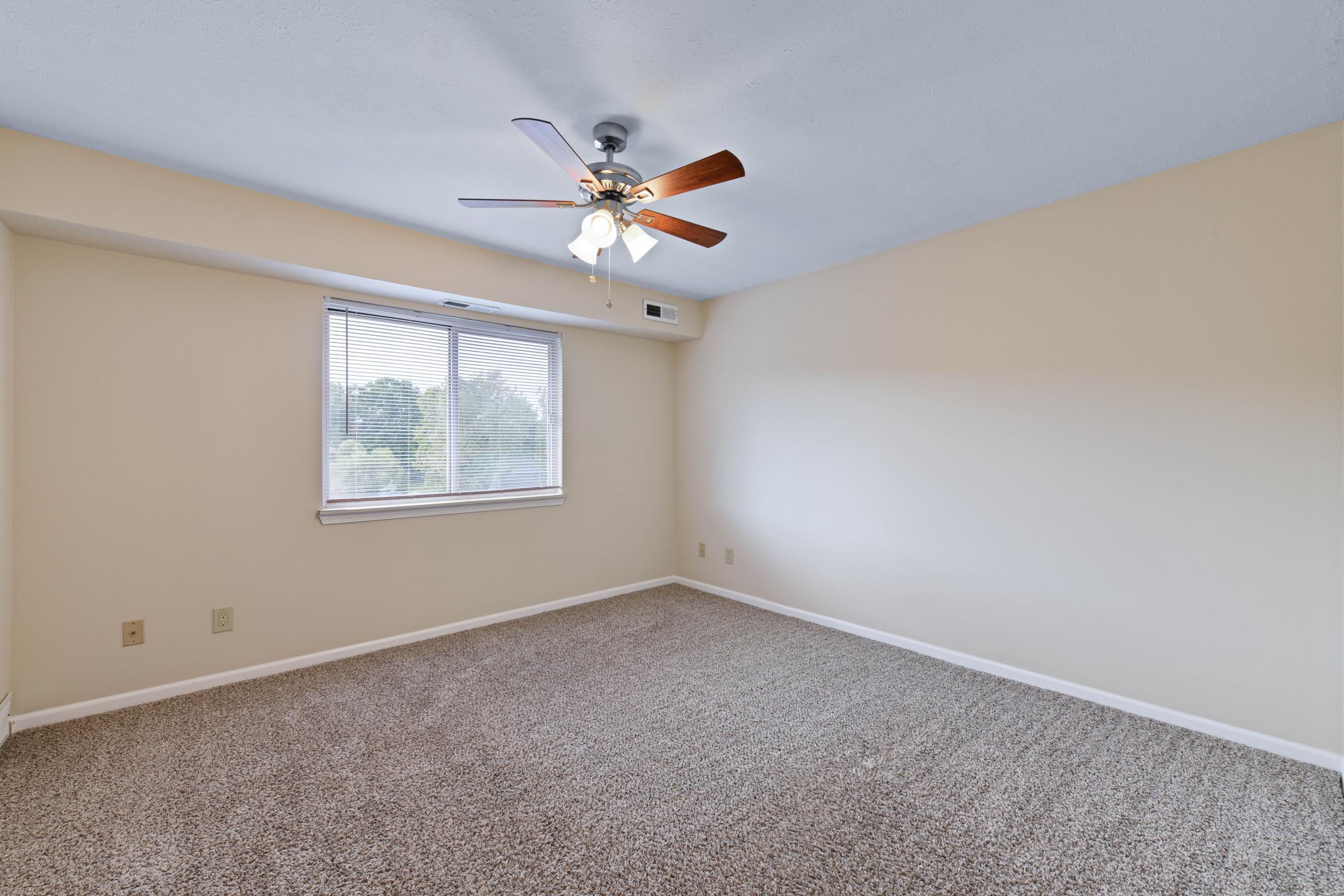 A spacious, vacant room with light beige walls and a ceiling fan featuring wooden blades. The floor is covered in light brown carpet. A window on the left allows natural light to enter, offering a view of greenery outside. The overall atmosphere is bright and inviting.