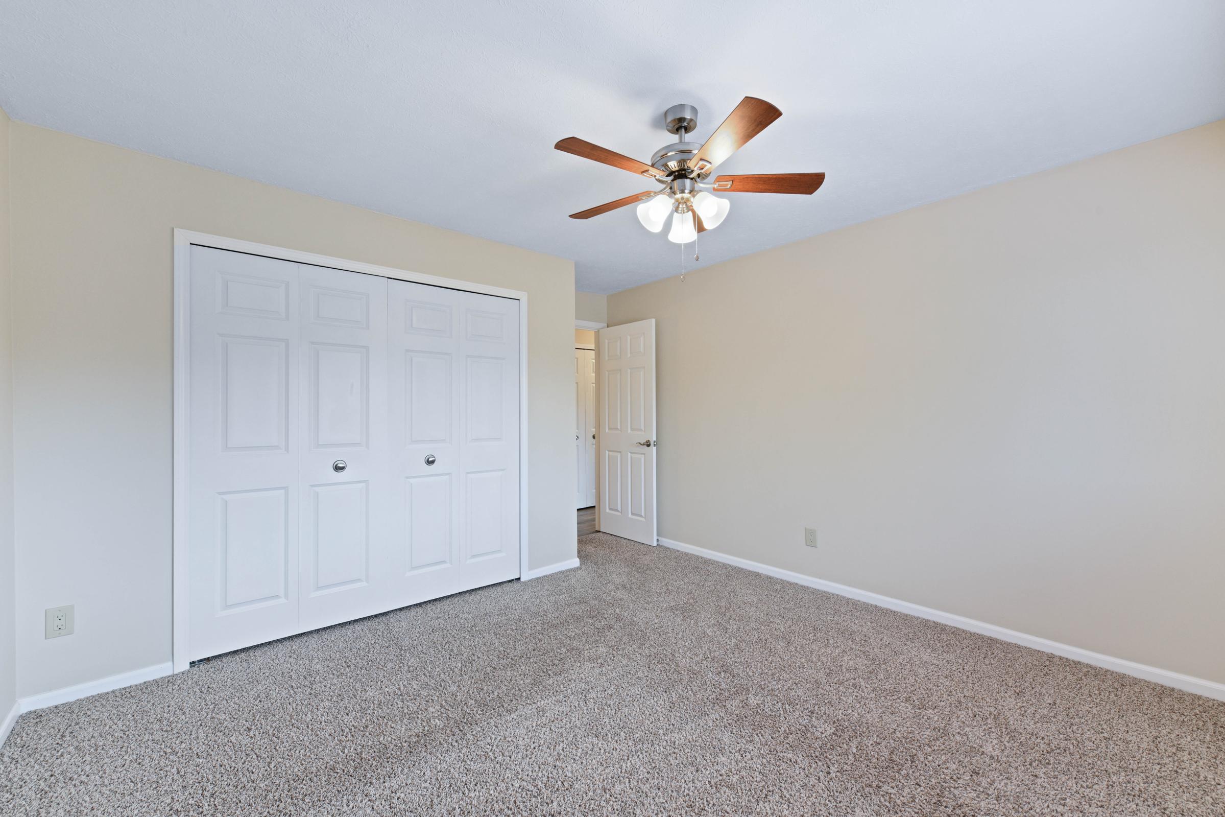 A spacious bedroom featuring light beige walls, a ceiling fan with wooden blades, and plush, neutral-colored carpeting. There are two sliding closet doors on one wall, and a doorway leading to another room in the background. The area is bright and well-lit.