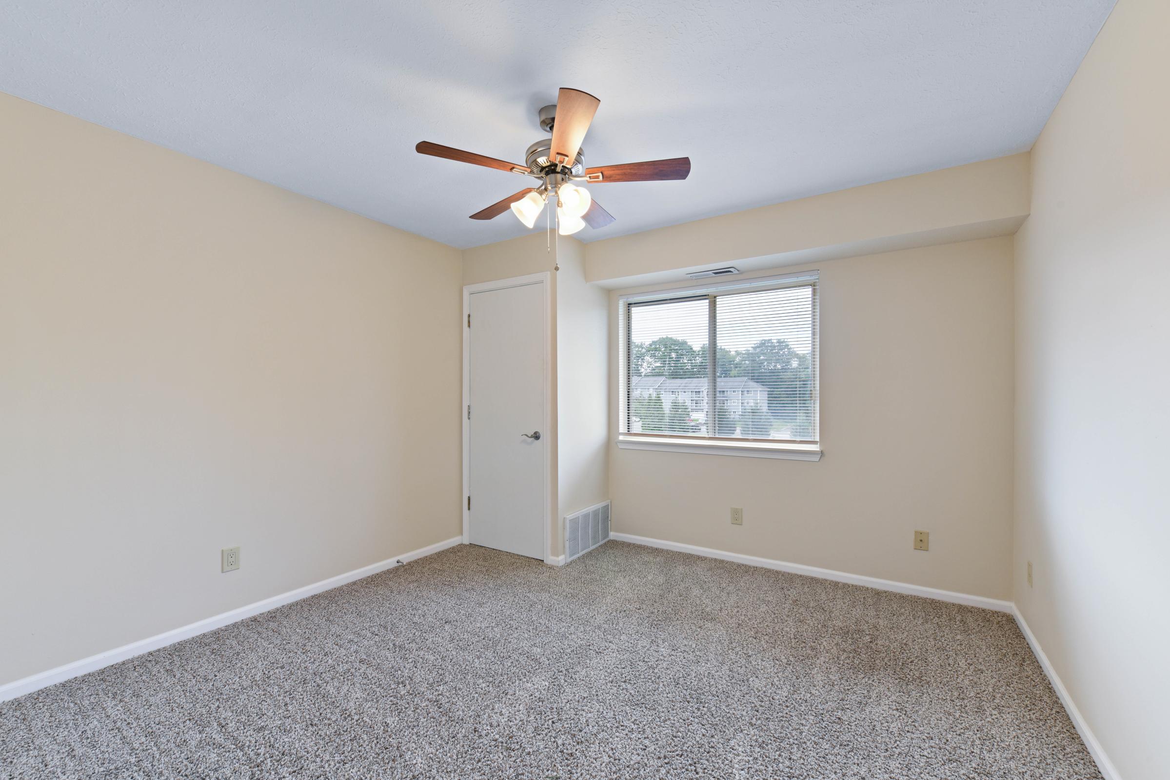 Empty room with beige walls and light brown carpet, featuring a ceiling fan with wooden blades. A window allows natural light to enter, and there is a simple door leading to another space. The overall atmosphere is bright and spacious, with no furniture present.