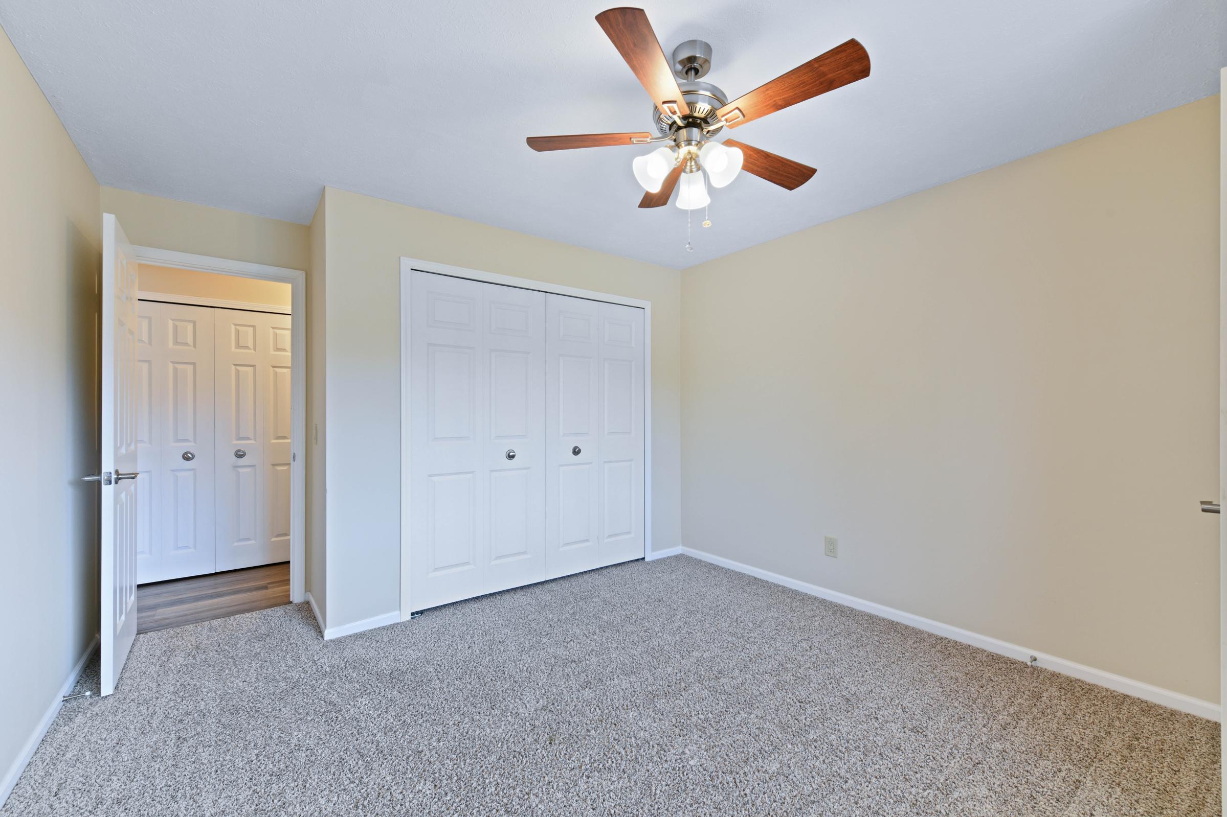 A spacious, light-colored bedroom featuring a ceiling fan with wooden blades. The room has beige walls and soft carpet flooring. A double closet with white sliding doors is visible on one side, and an open door leads to a hallway with additional closed doors, enhancing the room's inviting and airy feel.
