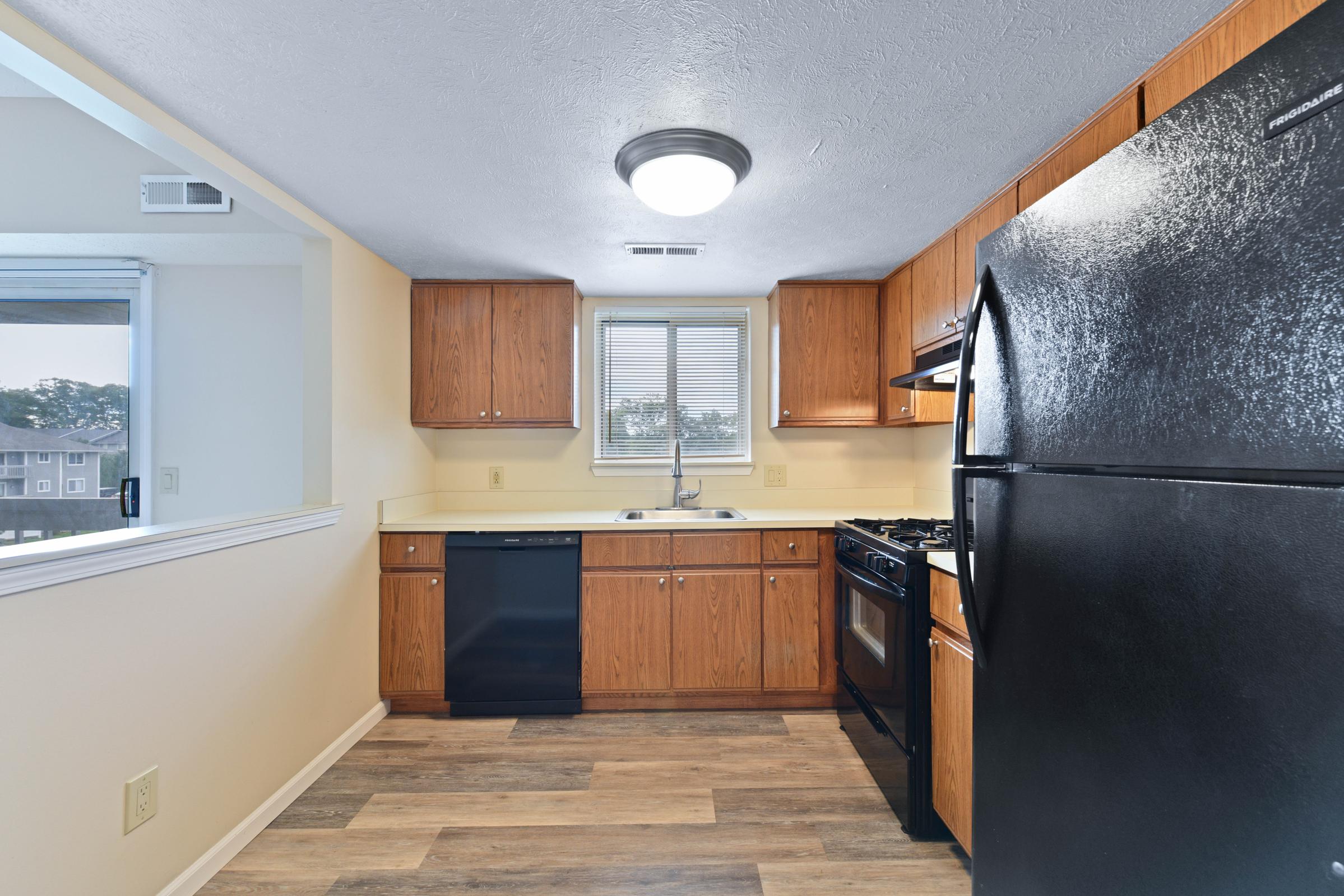 A modern kitchen featuring wooden cabinets, a black refrigerator, a built-in dishwasher, and a gas stove. The countertop has a sink with a window above it, providing natural light. The flooring consists of light wood-like tiles, and the walls are a neutral color.
