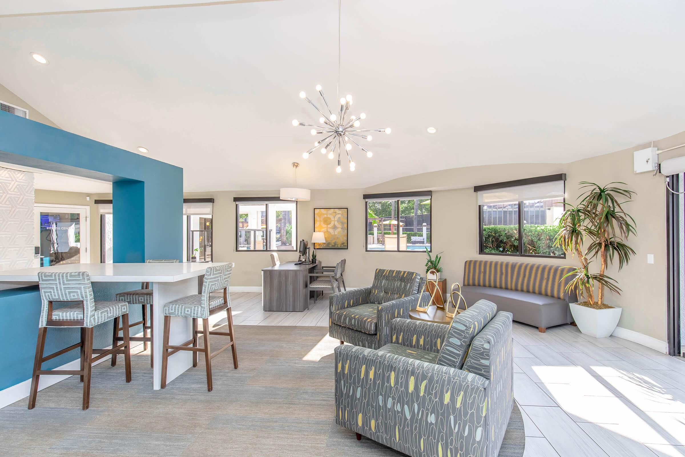 A modern lounge area featuring a seating arrangement with patterned armchairs and a sleek couch. A round dining table sits next to a bar with high stools. Large windows allow natural light, and a decorative plant adds a touch of greenery. The design is contemporary with a light color palette and stylish lighting fixture.