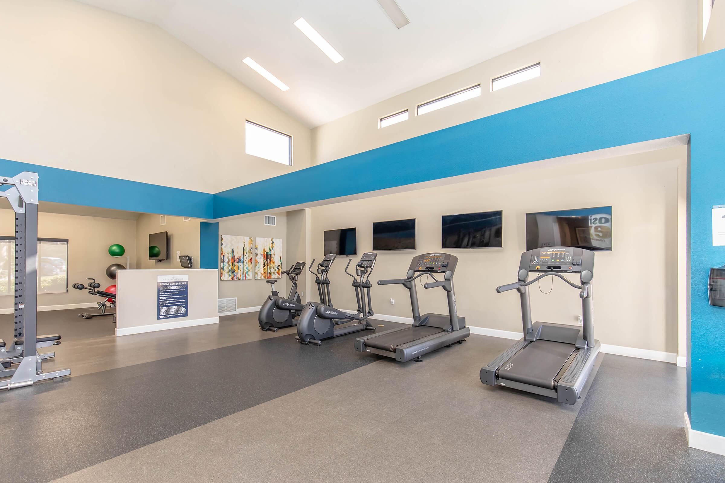 A modern fitness center featuring treadmills, exercise equipment, and large windows for natural light. The walls are painted in light colors with a blue accent, and there are several wall-mounted screens. The floor has a dark rubber surface, creating a welcoming workout environment.