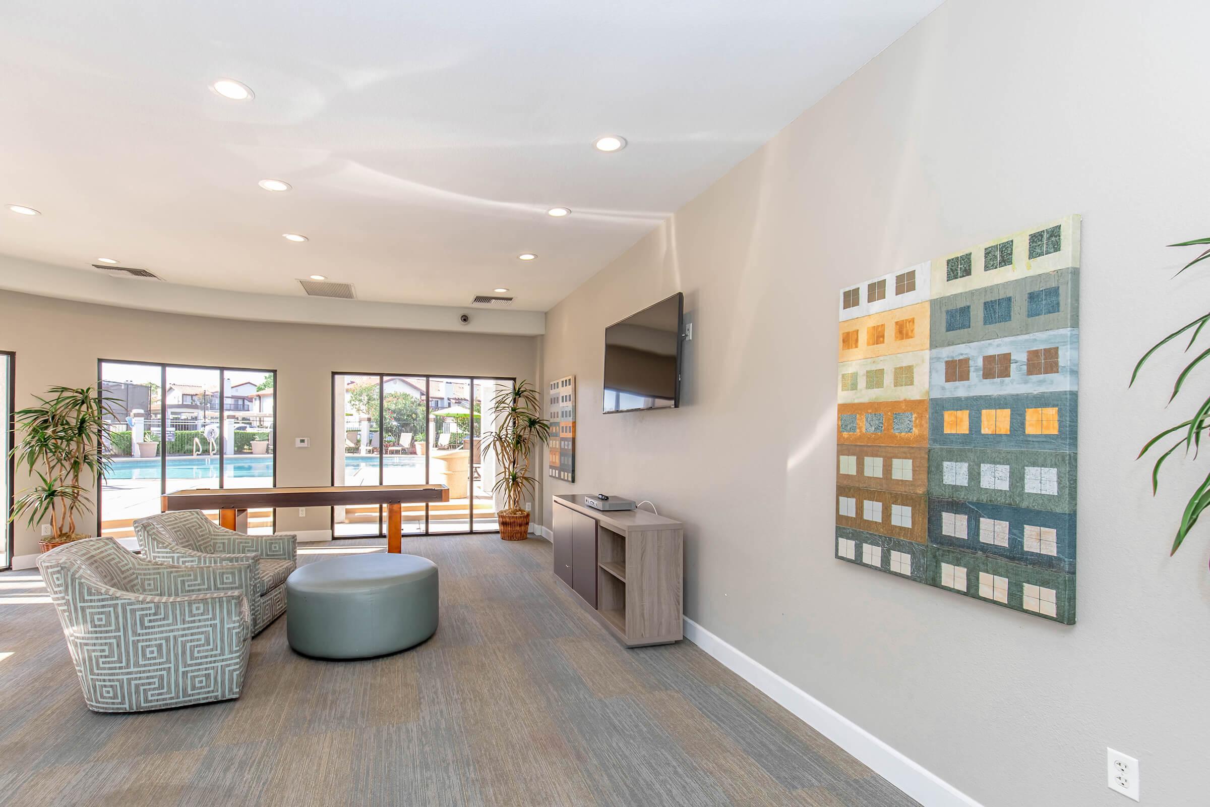 Modern interior of a lounge area featuring a light-colored wall with large windows showcasing a pool view. There is a gray fabric ottoman, a wooden console table, a TV mounted on the wall, and a colorful abstract wall art piece. Tall indoor plants add a touch of greenery to the space.