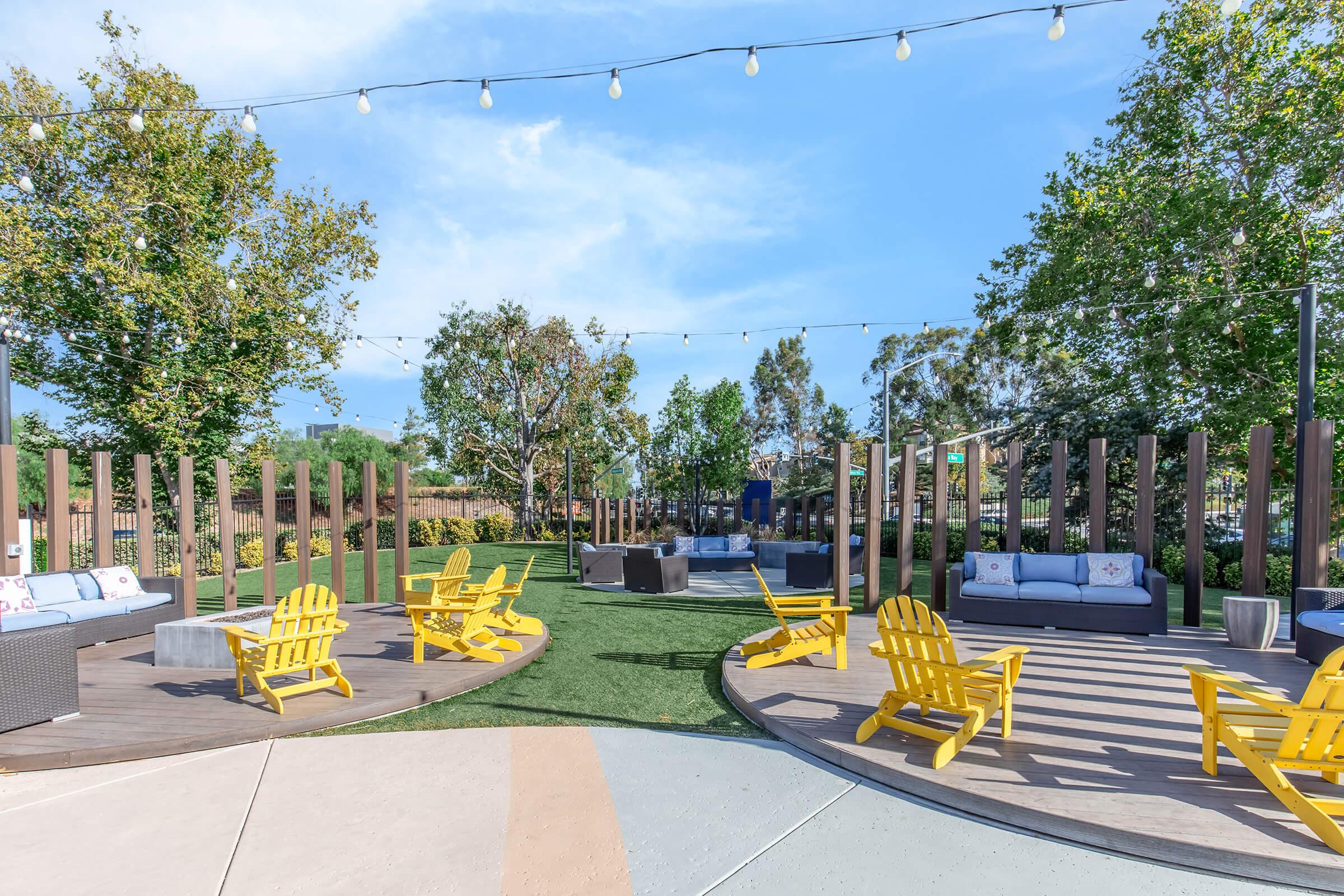 Outdoor seating area featuring yellow Adirondack chairs and gray couches on a wooden deck surrounded by lush greenery and trees. String lights hang above, creating a warm atmosphere. Perfect for relaxation and social gatherings in a serene environment.