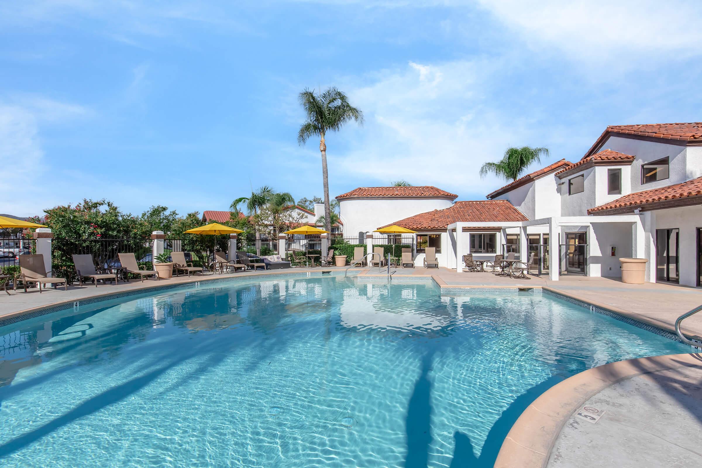 A sunny outdoor pool area featuring a curved swimming pool surrounded by lounge chairs, palm trees, and umbrellas. There are modern buildings in a Mediterranean style in the background, with clear blue skies overhead, creating a relaxing atmosphere.