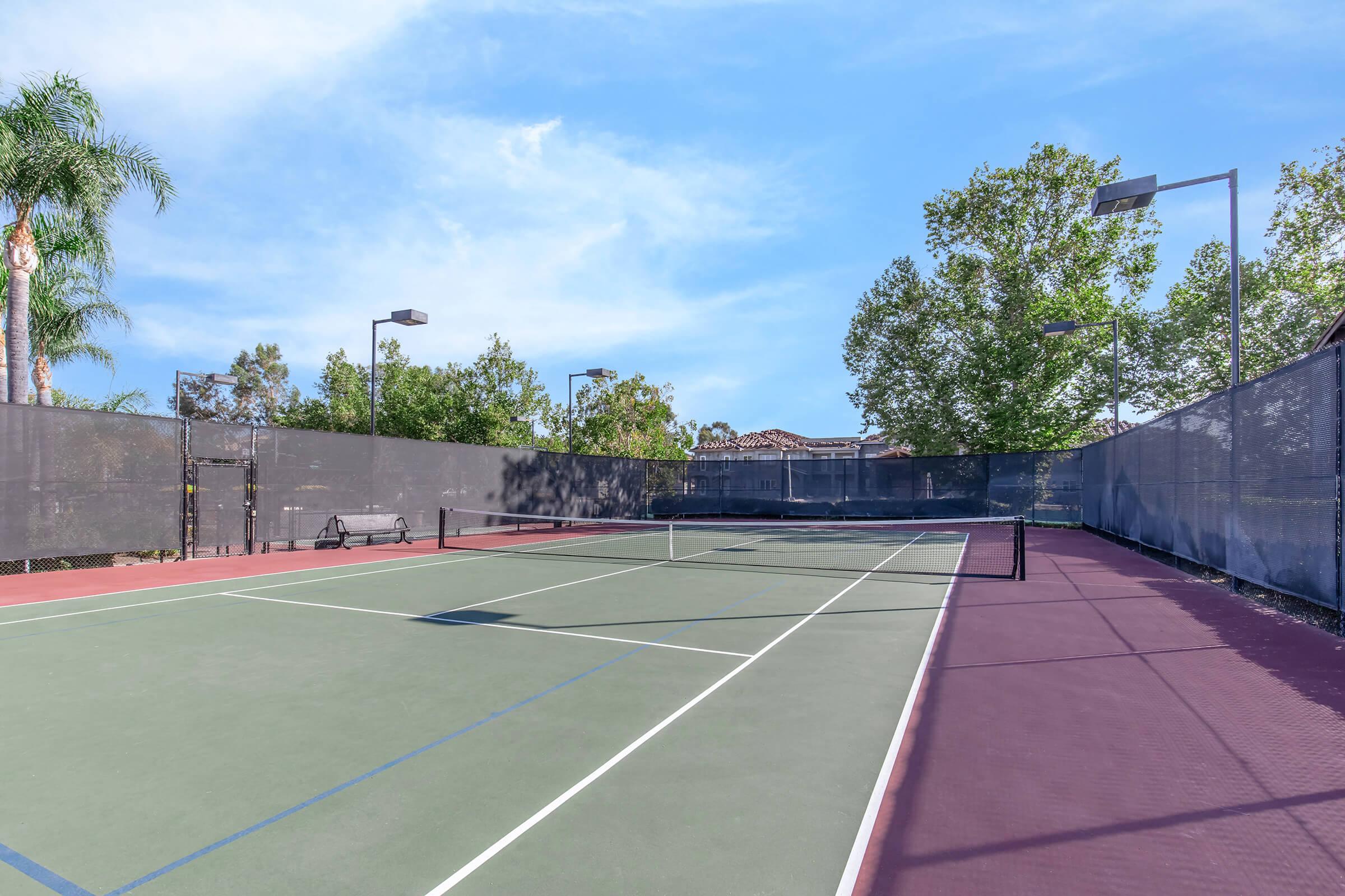 Empty tennis court with a green playing surface and a purple area, surrounded by tall trees and a black fence. The sky is clear with a few clouds, and there are benches and lighting fixtures visible.