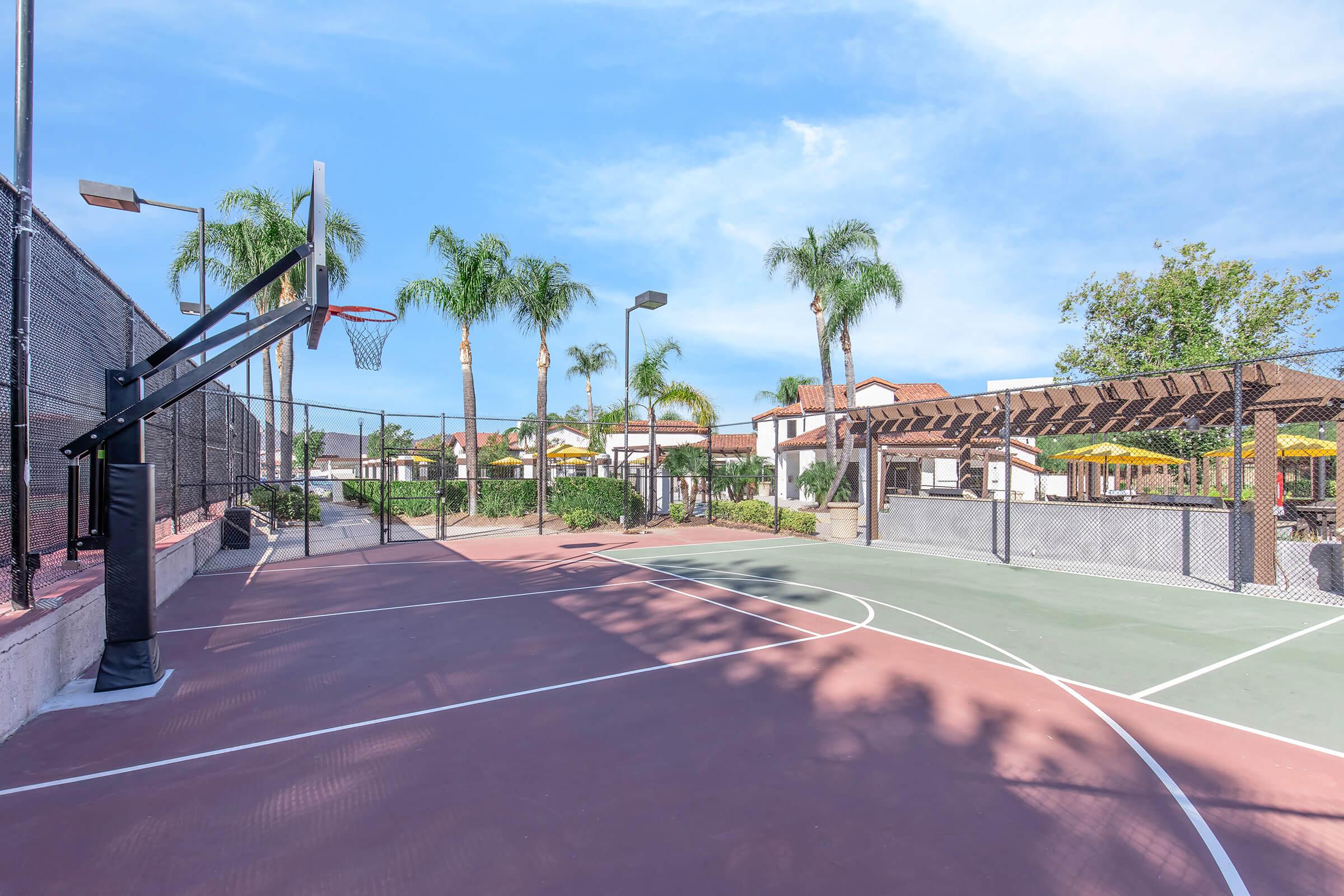 A basketball court featuring a freestanding hoop, surrounded by palm trees. The court has two different surface colors, with a fence enclosing the area. In the background, there are structures and umbrellas, suggesting a recreational facility or park setting under a clear blue sky.