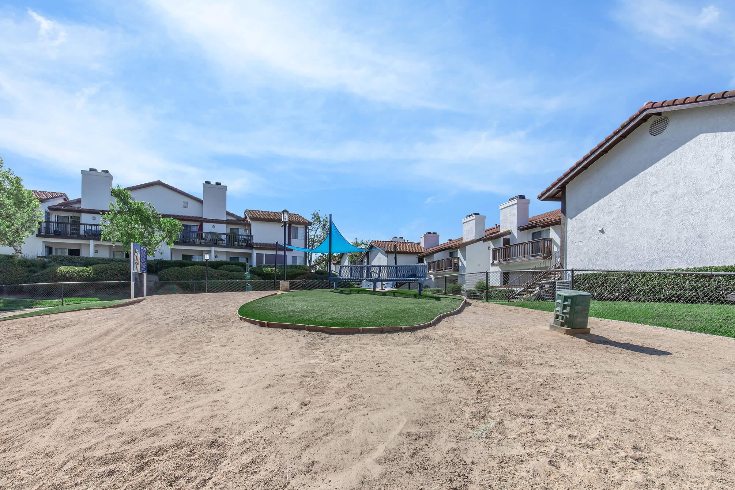 A playground area with a sandy surface and green grass, surrounded by residential buildings with balconies. The sky is clear with a few clouds, and there are trees and landscaping visible in the background.
