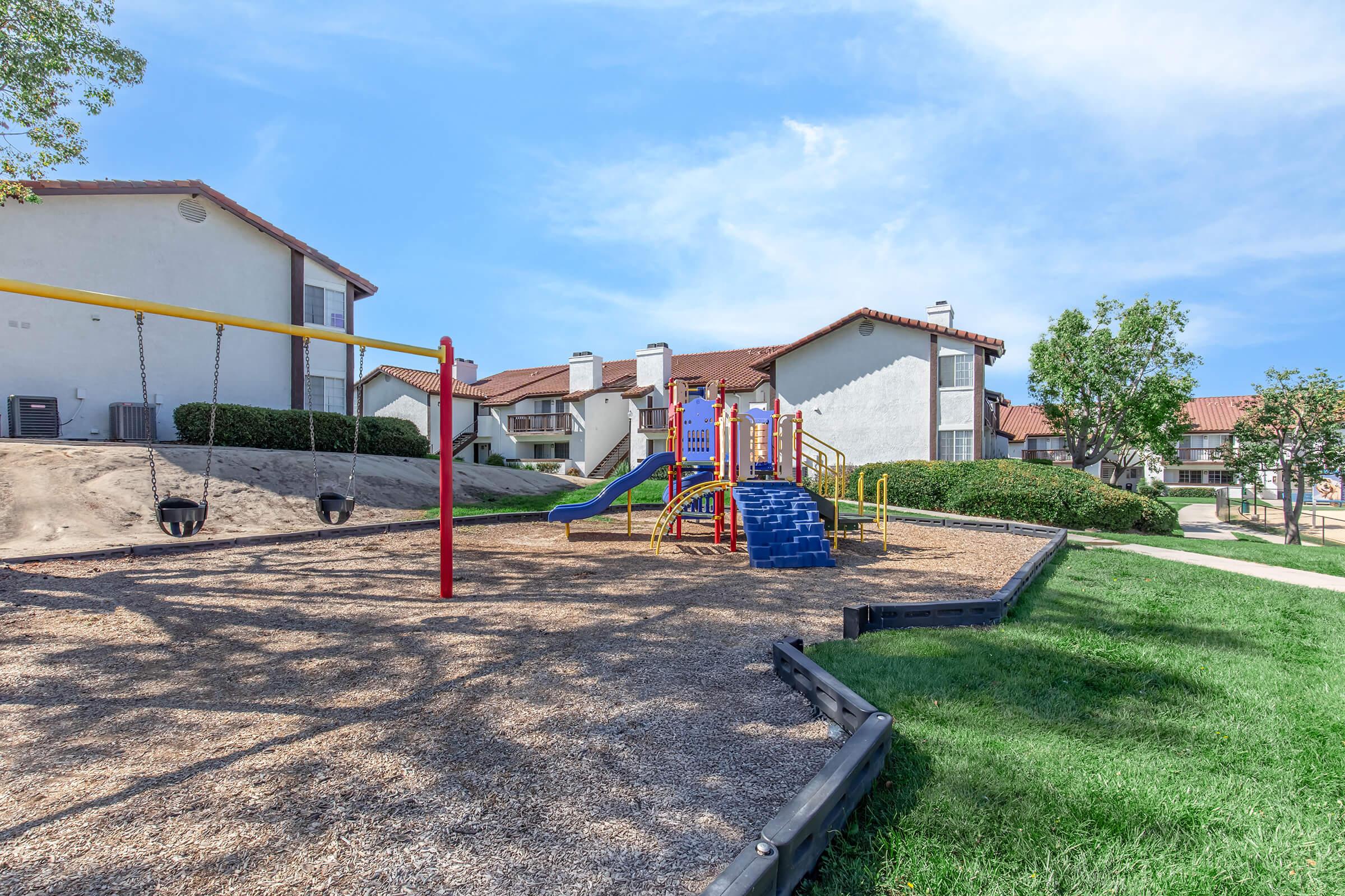 A colorful playground featuring a slide and climbing structure, alongside swings, situated on a sandy area. Lush green grass surrounds the playground, with residential buildings in the background and a clear blue sky overhead.