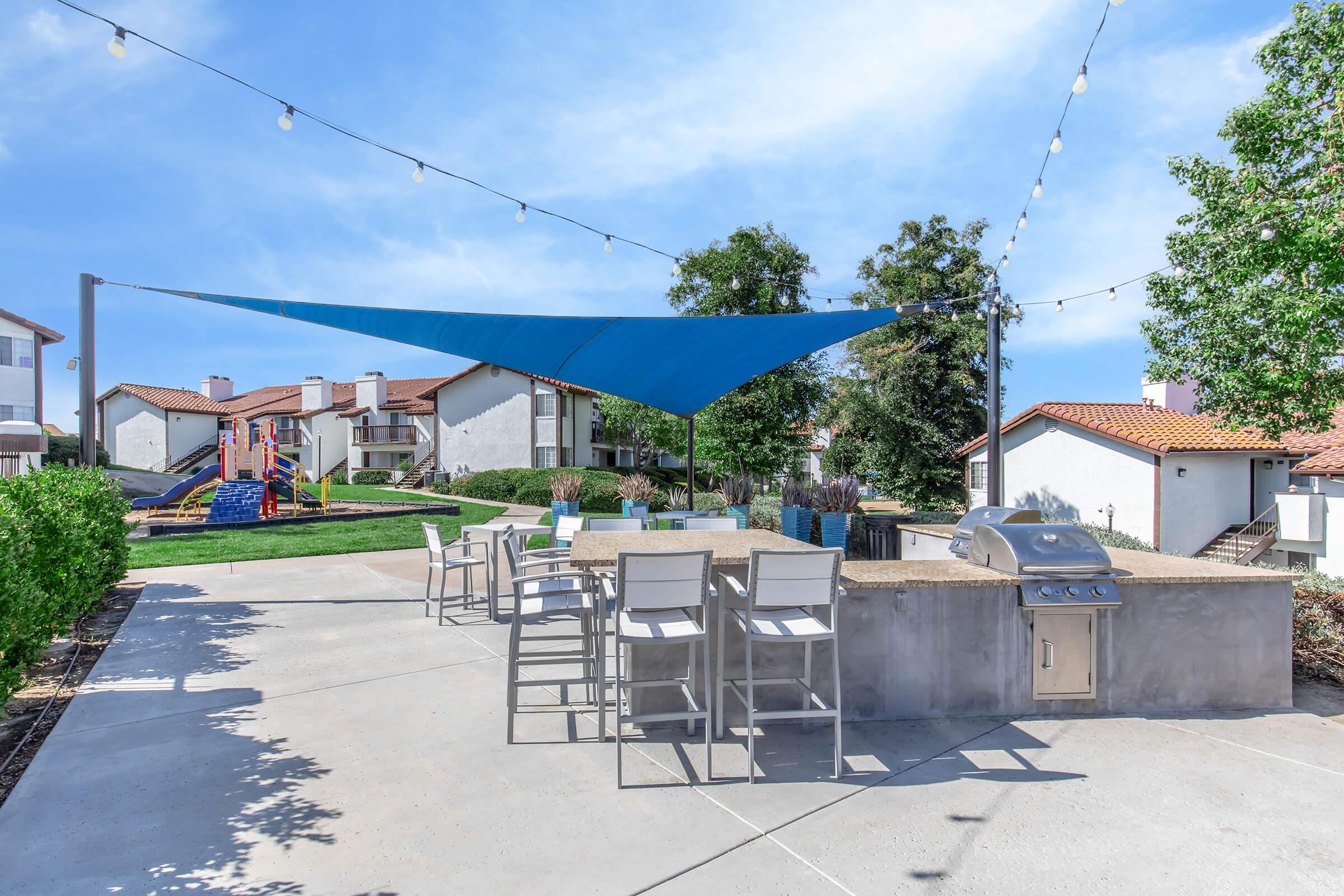 Outdoor common area featuring a shaded seating area with bar stools, a barbecue grill, and a blue shade canopy. Lush greenery surrounds the space, with residential buildings in the background. String lights are hung above, creating a welcoming atmosphere. Ideal for gatherings and relaxation.