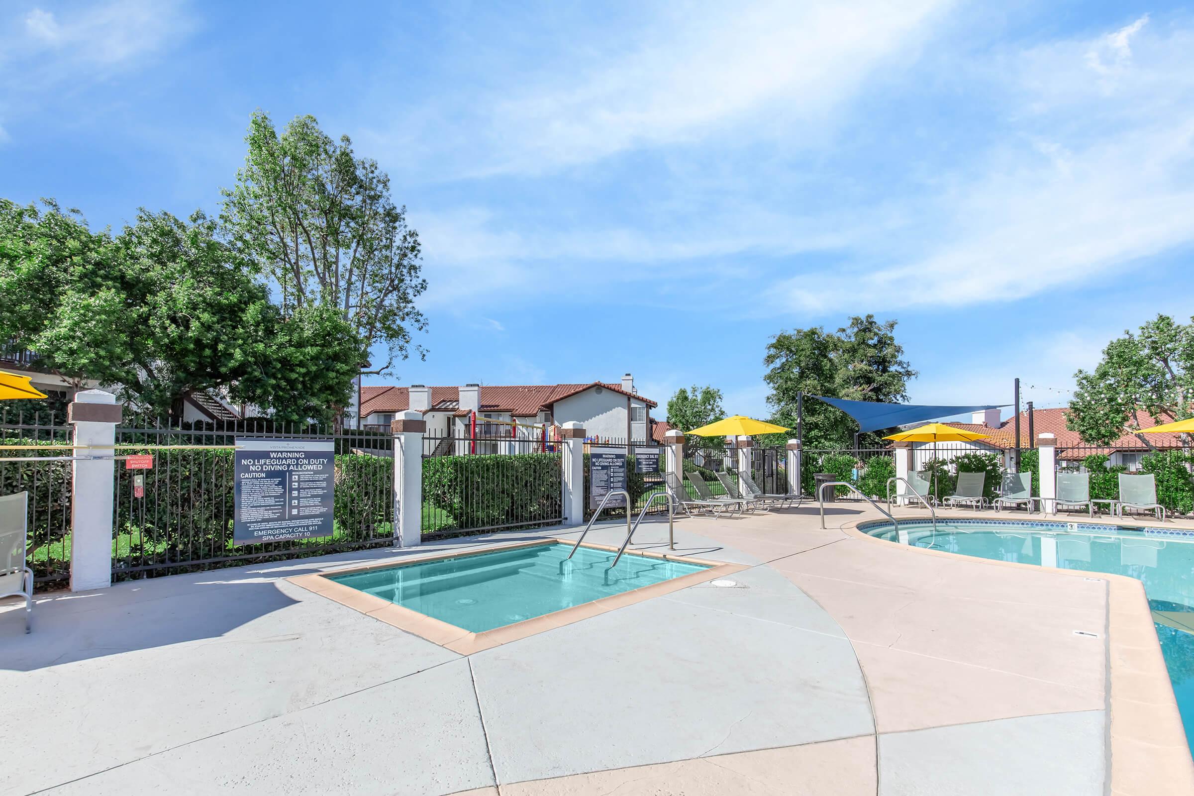 A well-maintained outdoor swimming pool area featuring a large main pool, a smaller spa, and several yellow umbrellas for shade. Surrounding greenery and fencing create a cozy atmosphere, with clear blue skies visible above. Informational signs are posted nearby.