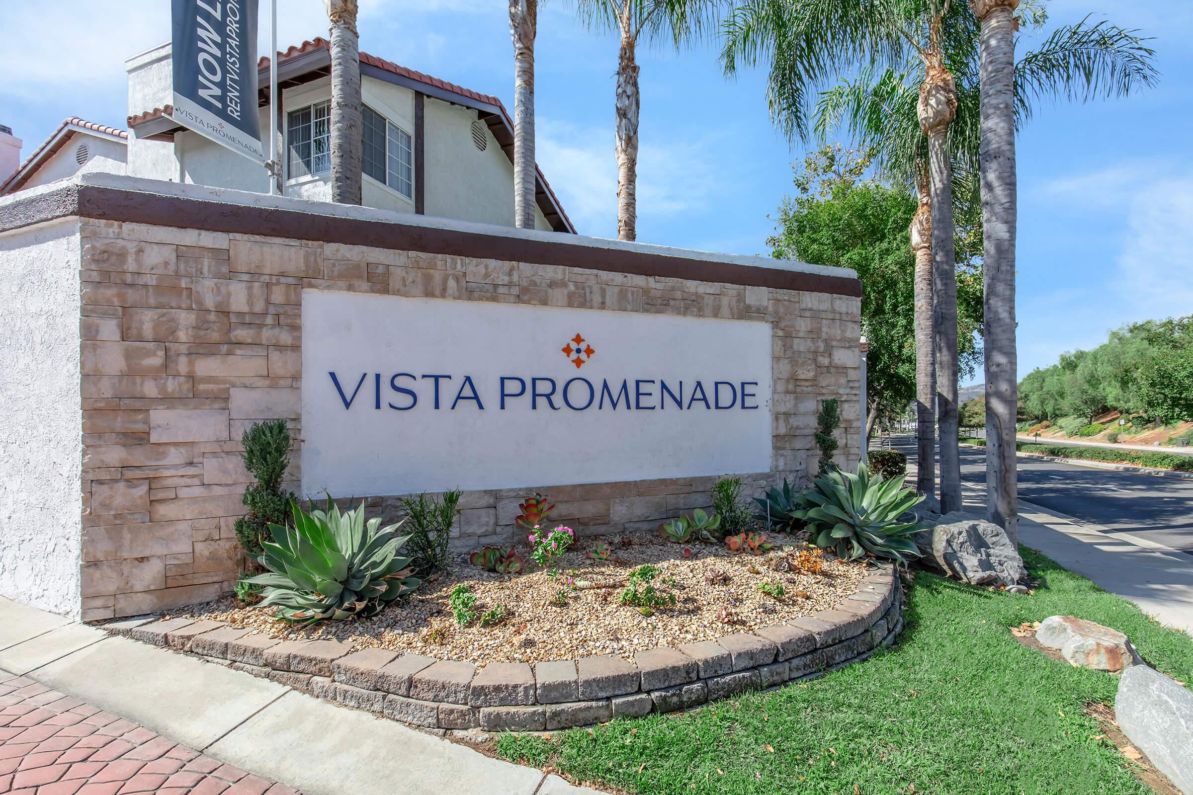 Sign for "Vista Promenade" displayed prominently in a landscaped area featuring a stone base, surrounded by decorative plants and rocks, with palm trees in the background and a clear blue sky overhead.