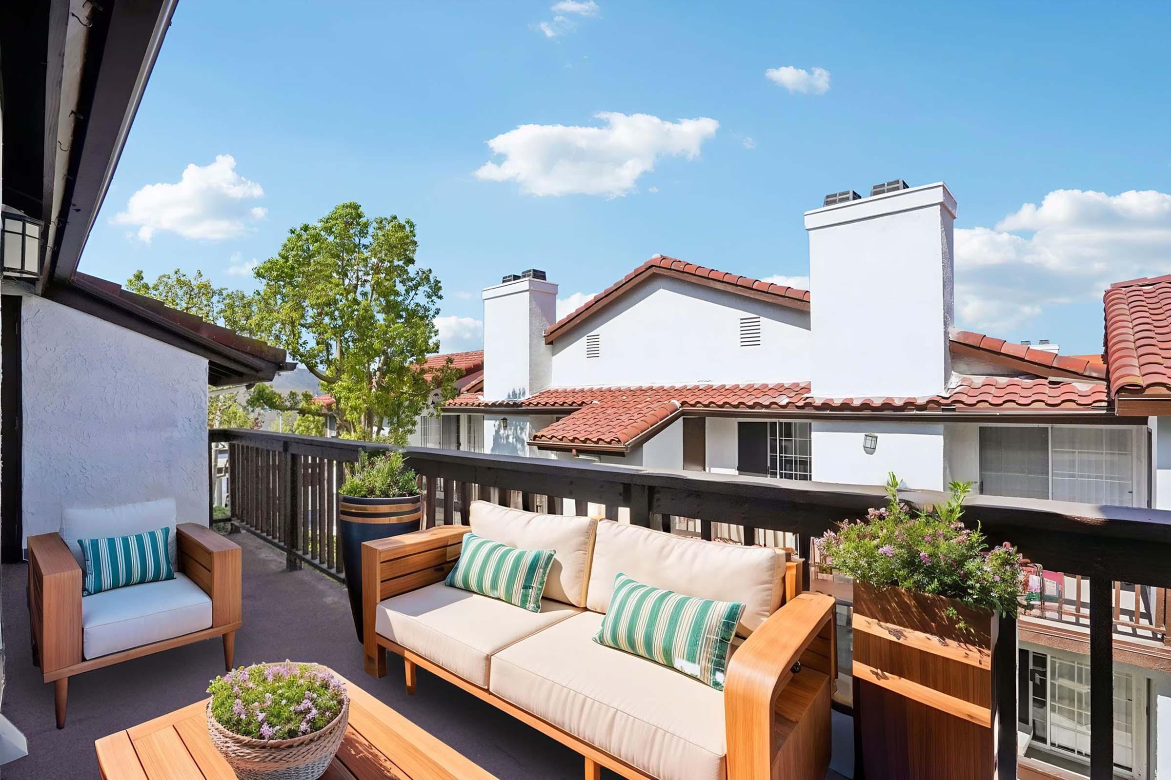 A cozy balcony featuring a wooden sofa with striped cushions and a wooden coffee table. Potted plants add greenery, and the view includes rooftops and a clear blue sky with fluffy clouds. The setting exudes a relaxing, inviting atmosphere perfect for outdoor lounging.