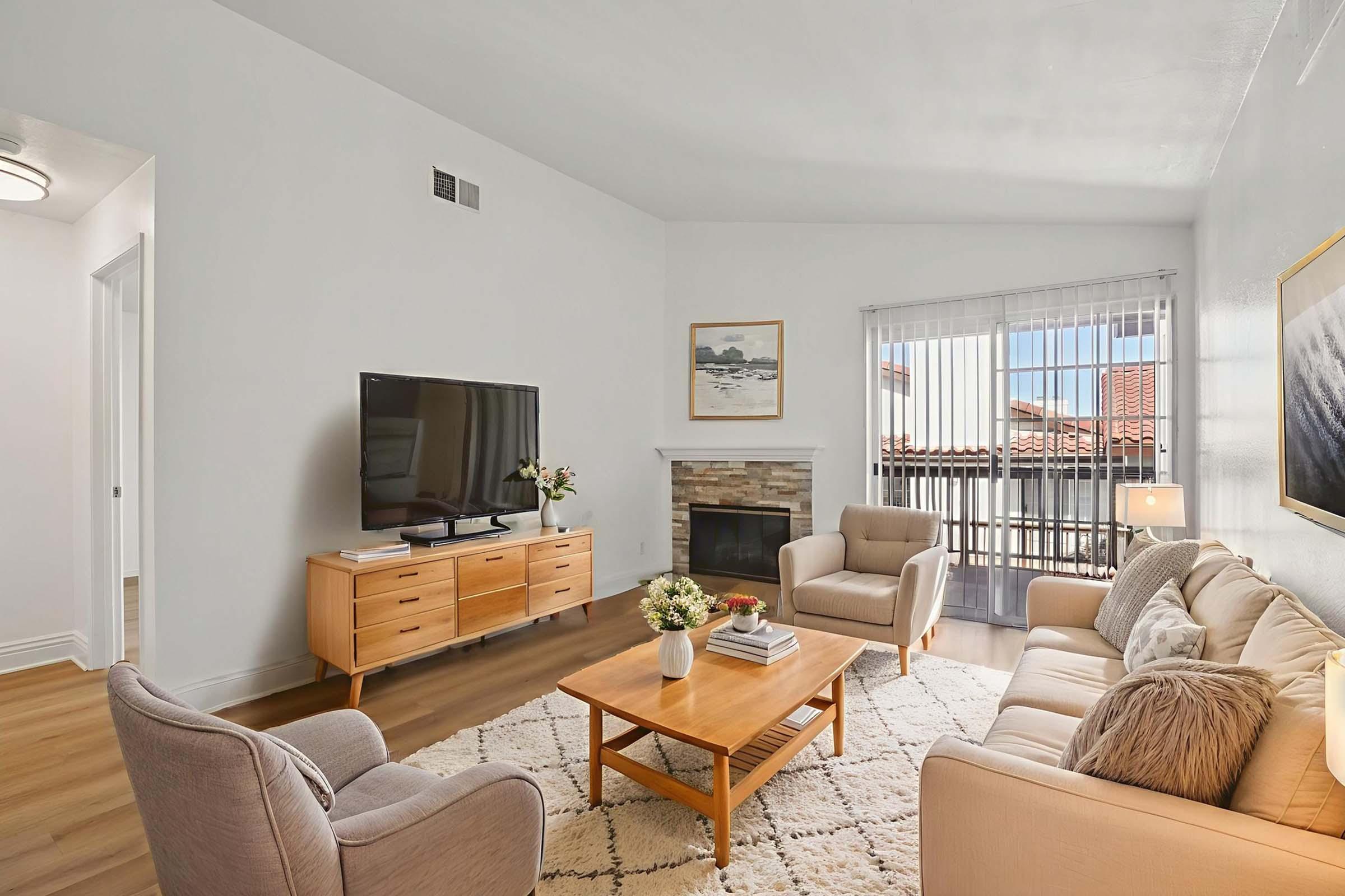 A bright and inviting living room featuring two beige sofas, a wooden coffee table, and a TV stand. A flat-screen TV is mounted on the wall, while a stone fireplace adds a cozy touch. Large windows with blinds provide natural light, and a piece of art hangs on the wall, enhancing the warm atmosphere.