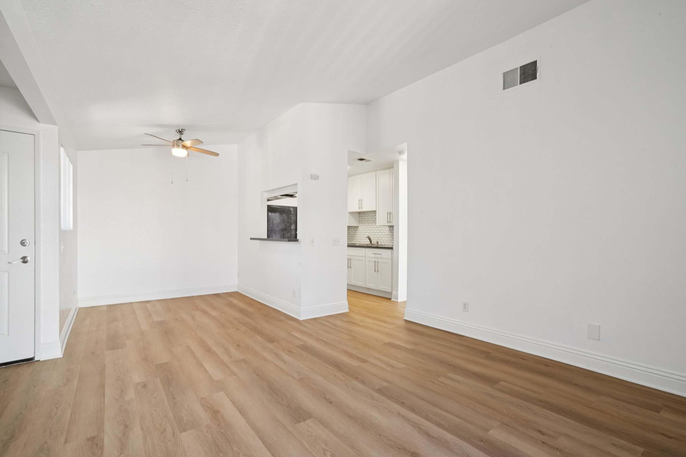 A spacious, minimalist living area featuring light wood flooring, a ceiling fan, and a white wall. The adjacent open kitchen is partially visible, showcasing modern cabinetry and a tiled backsplash. Natural light brightens the room, creating a welcoming atmosphere.