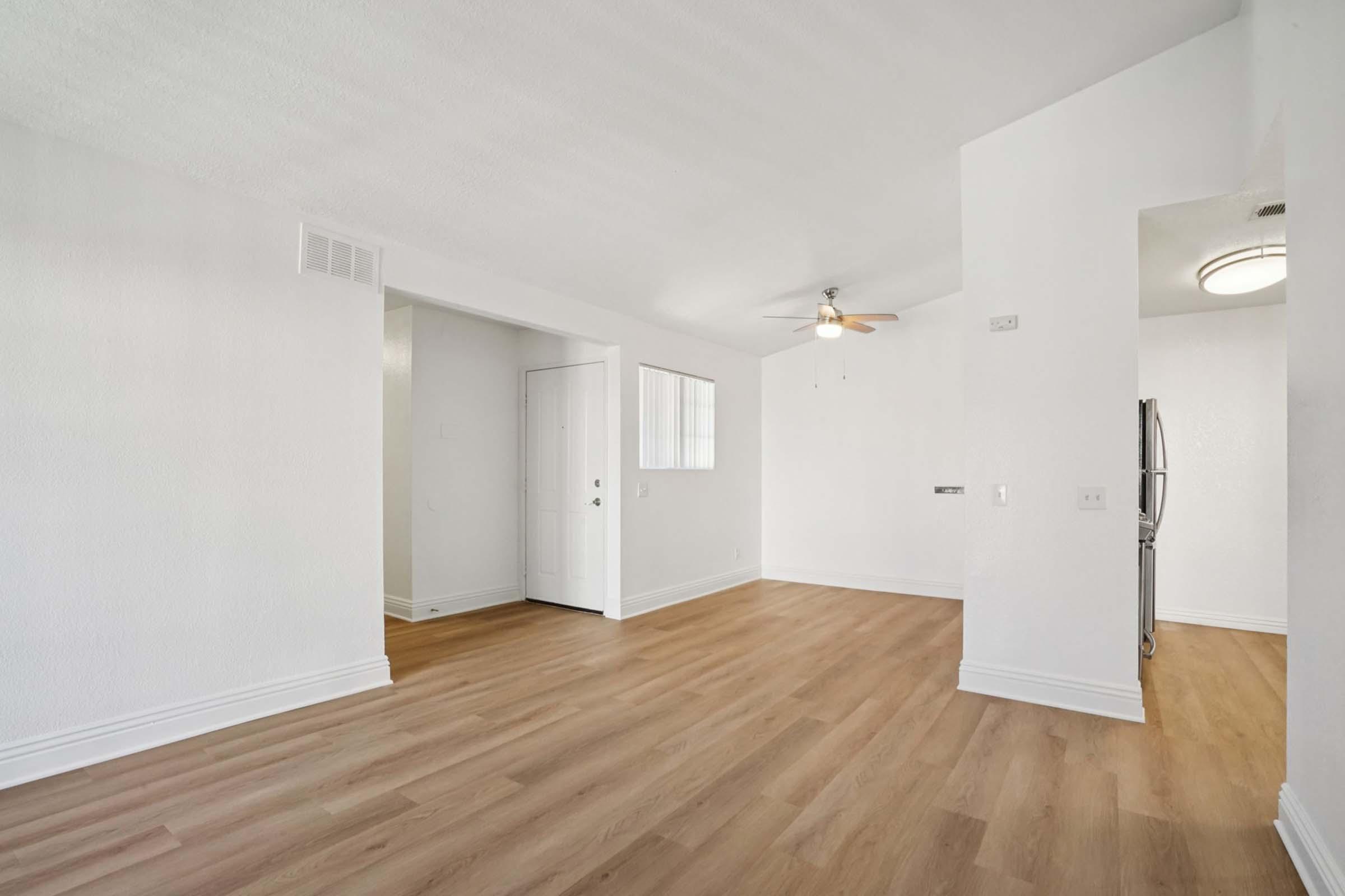 A spacious, empty room with light-colored walls and a wooden floor. A ceiling fan is installed, and there are windows allowing natural light. A door leads outside, and a corner features a silver refrigerator. The space appears well-maintained and ready for furnishing.