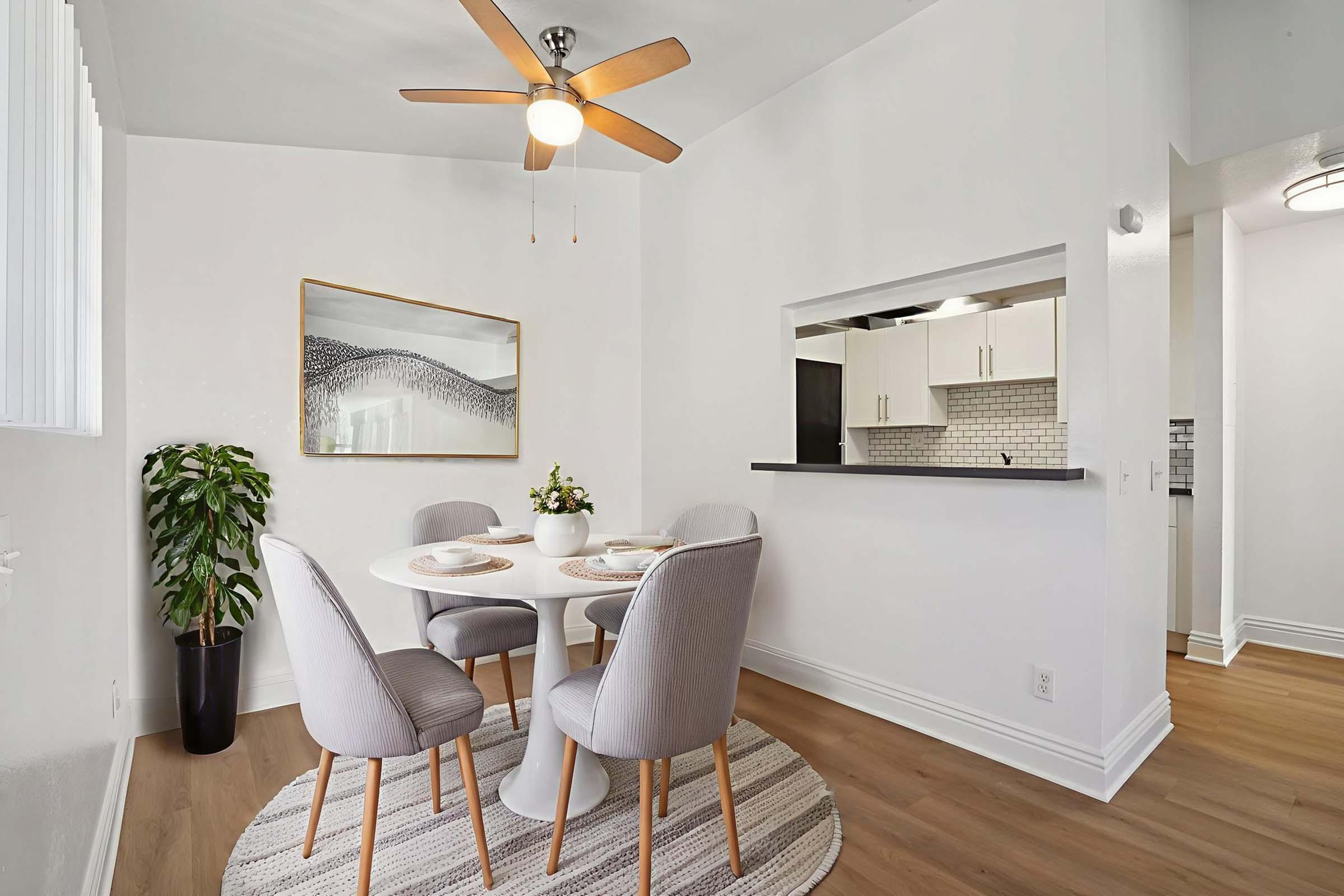A modern dining area featuring a round white table with four gray chairs, set with plates and decorative items. A ceiling fan hangs above, and there's a potted plant in the corner. In the background, an open kitchen area with sleek cabinetry and stylish backsplash is visible, enhancing the contemporary feel of the space.