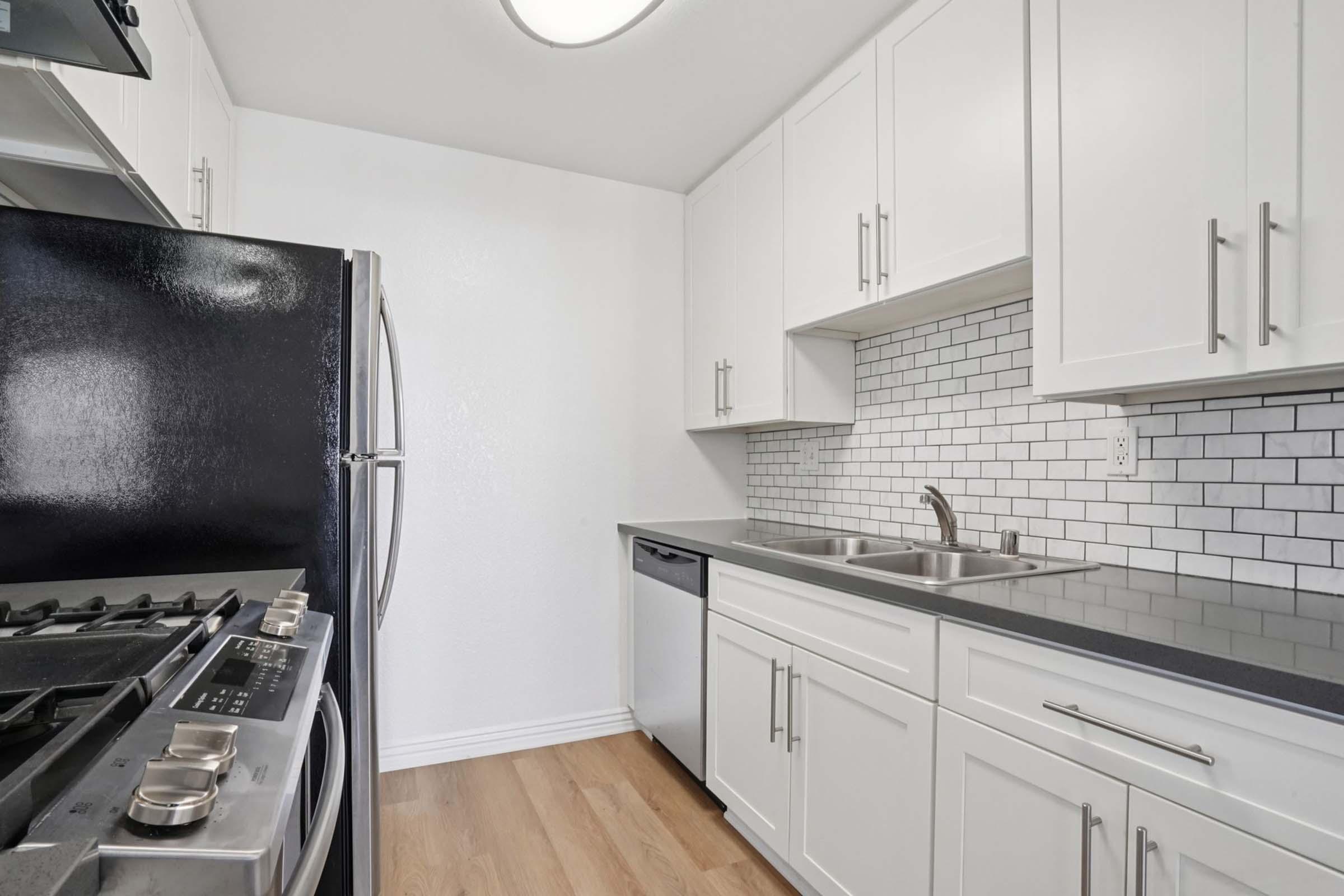 Modern kitchen featuring white cabinets, a black refrigerator, stainless steel appliances, and a two-basin stainless steel sink. The backsplash consists of white subway tiles. The countertop is dark, and the floor is light hardwood. Overall, the design emphasizes a clean and contemporary aesthetic.