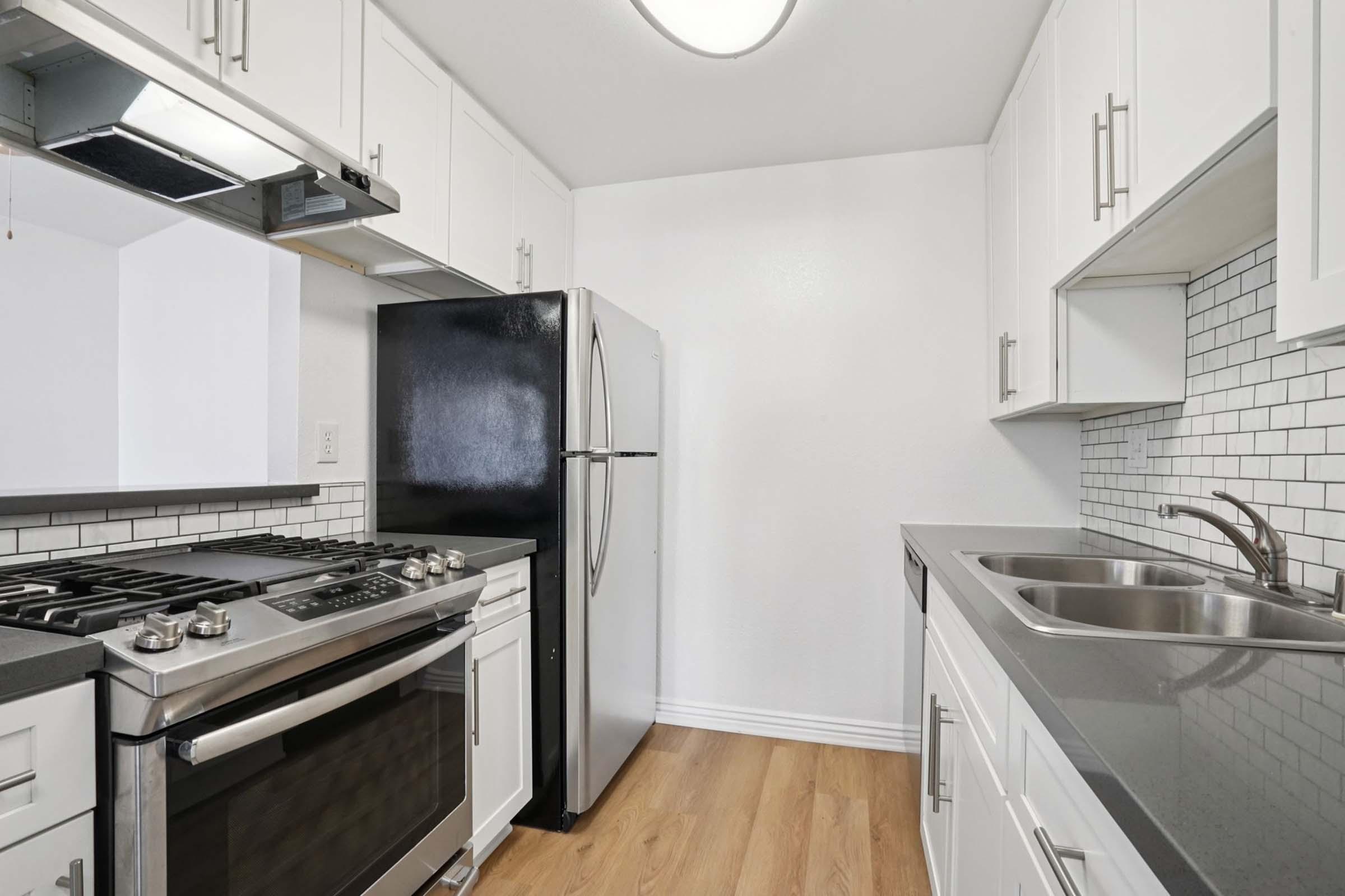 Modern kitchen with white cabinetry, stainless steel appliances, and black countertops. Features a gas stove, double sink, and a black refrigerator. The backsplash is tiled in a white square pattern. Wooden flooring complements the contemporary design.
