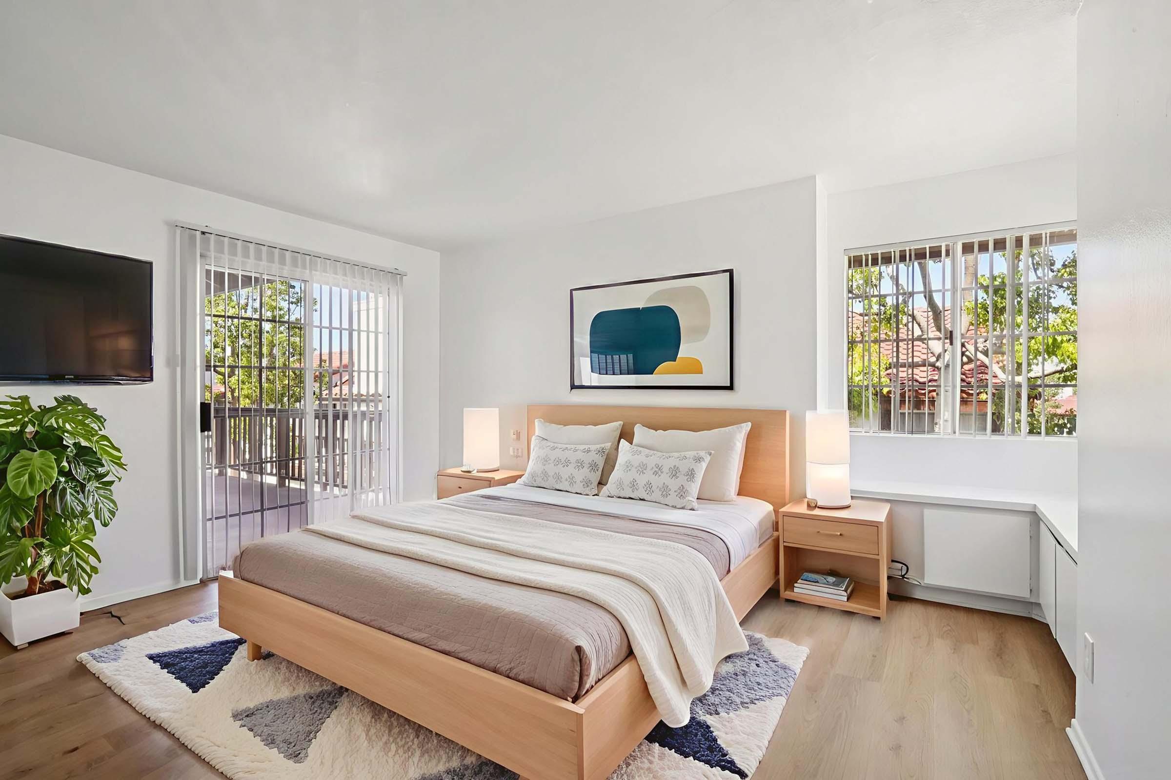 A bright and modern bedroom featuring a wooden bed with a light gray blanket, two bedside lamps, and a large piece of abstract art on the wall. There are windows with blinds allowing natural light, a TV mounted on the wall, and a potted plant, creating a cozy and inviting atmosphere.