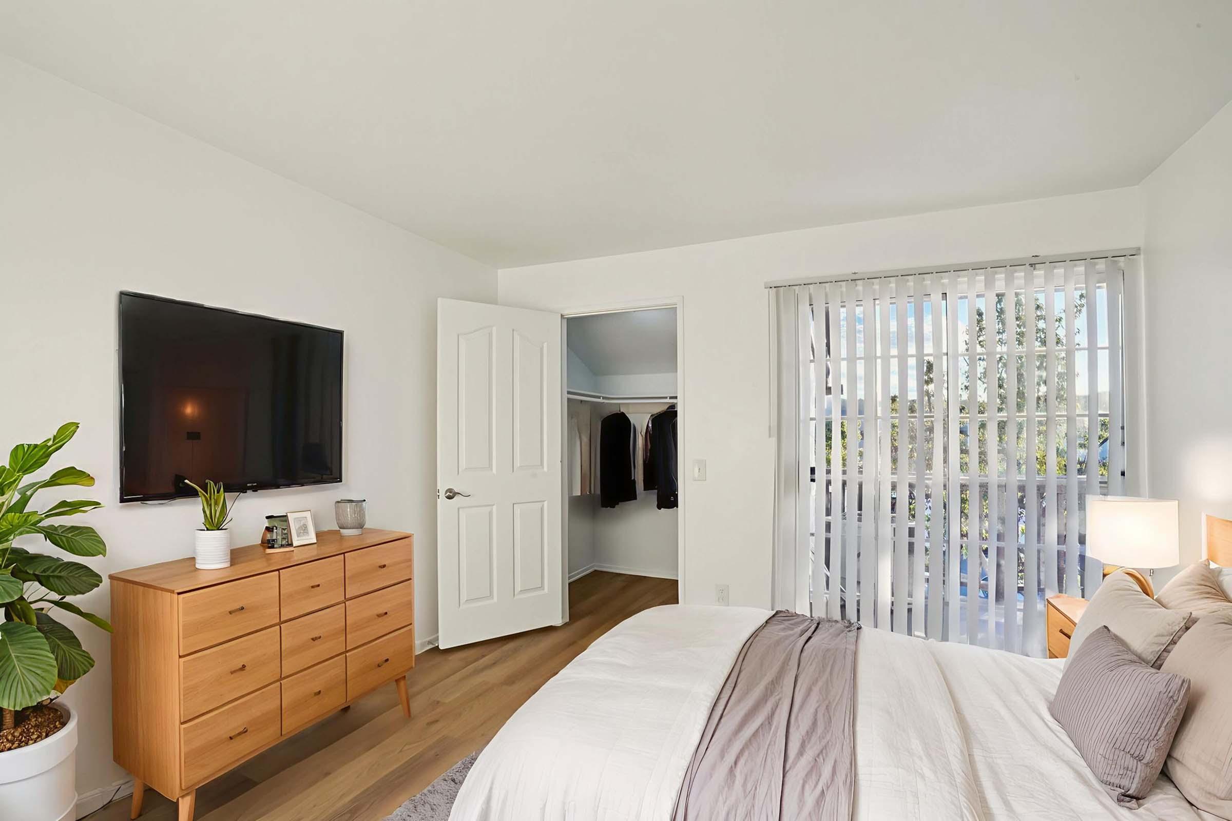 A modern bedroom featuring a comfortable bed with neutral bedding and a decorative throw. There's a wooden dresser against the wall, a flat-screen TV mounted above it, and a potted plant. A door leads to a closet, and large sliding glass doors allow natural light to enter the room, revealing a view outside.