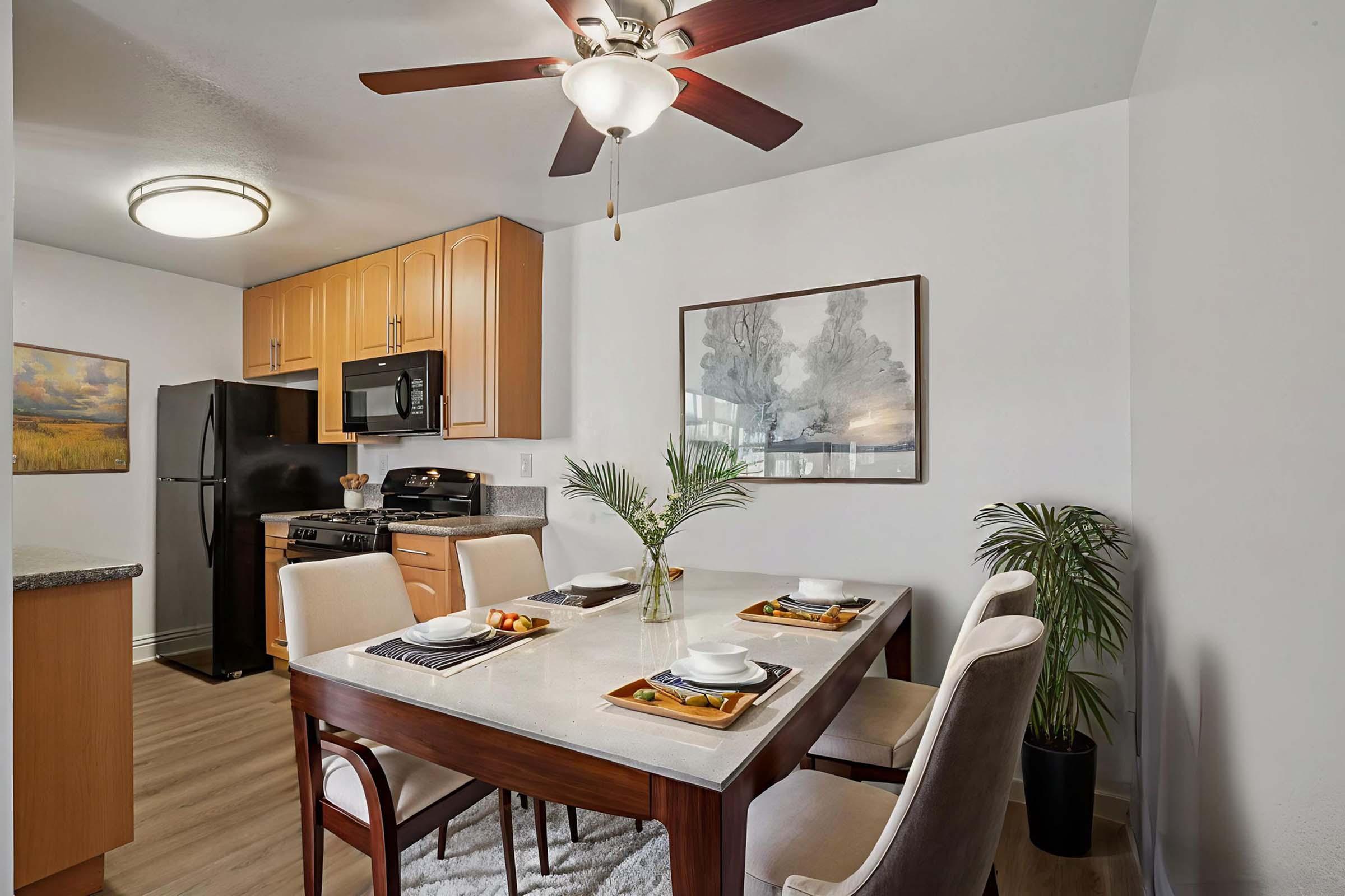 A modern kitchen and dining area featuring a wooden table set for four with plates and silverware. In the background, there's a refrigerator, black stove, and wooden cabinets. A ceiling fan hangs above, and potted plants add a touch of greenery. Bright artwork decorates the walls, enhancing a cozy ambiance.