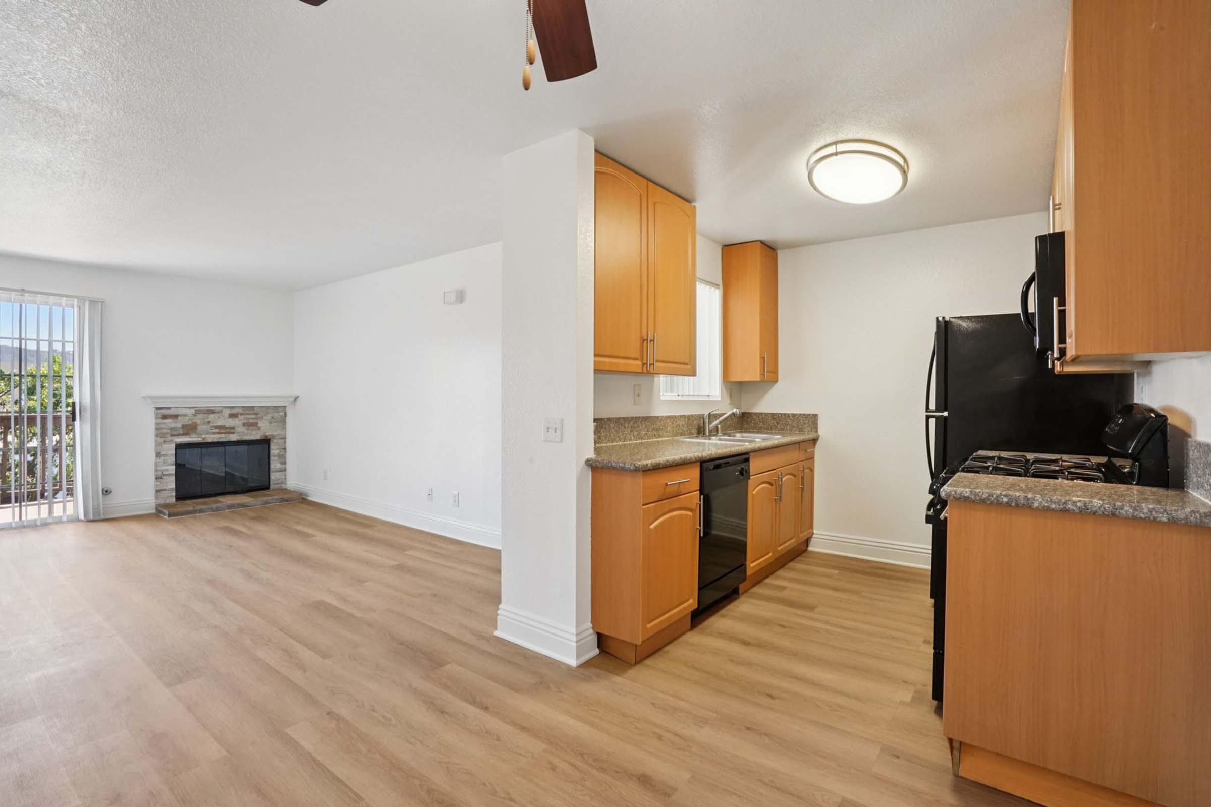 A modern open-concept living space featuring a kitchen with wooden cabinetry and black appliances. To the left, a cozy living area with a stone fireplace and large windows allowing natural light. The flooring is light-colored laminate, enhancing the spacious feel of the room.