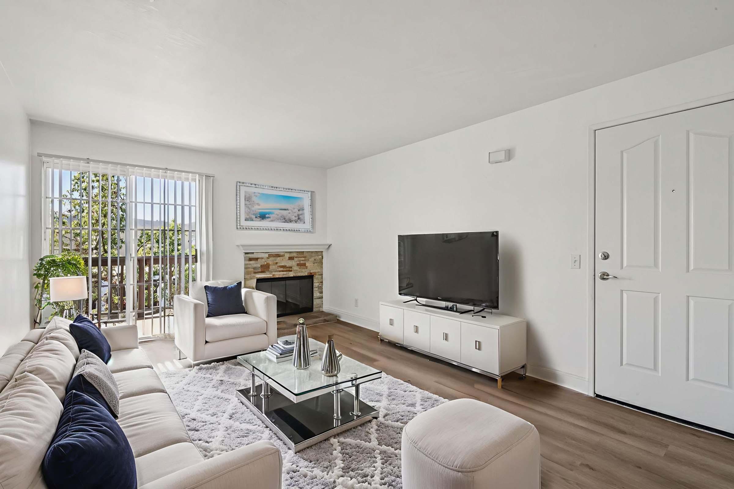 Modern living room featuring a neutral color scheme with a white sofa and armchairs, a glass coffee table on a plush rug, a stone fireplace, and a wall-mounted TV. Natural light streams through sliding glass doors with a balcony view. Decorative elements include plants and wall art, creating a cozy atmosphere.