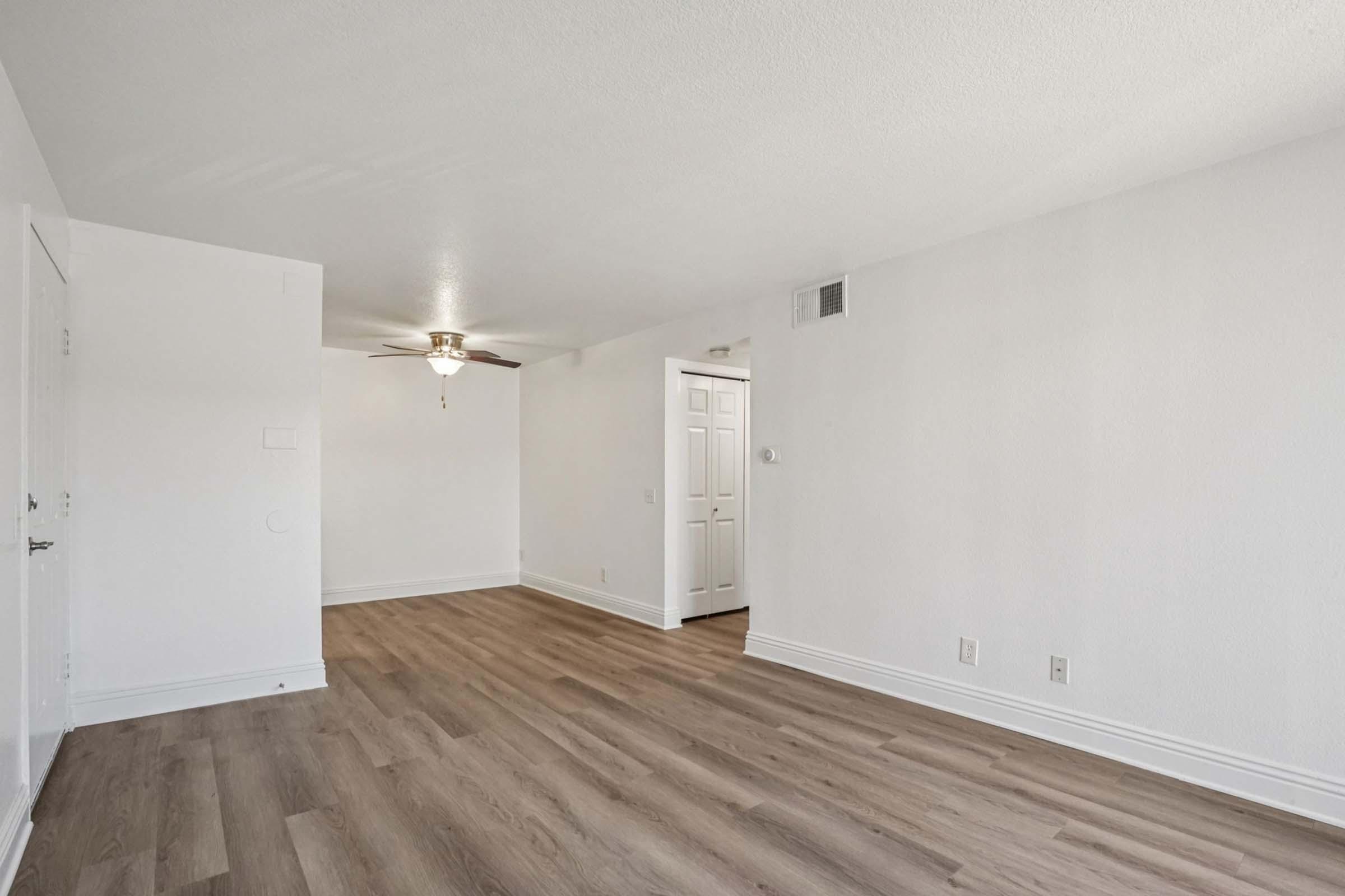 A bright, empty living room featuring light-colored walls and a ceiling fan. The floor is finished with light wood laminate. A doorway leads to another room, and the overall space is clean and well-lit, ready for furnishings.