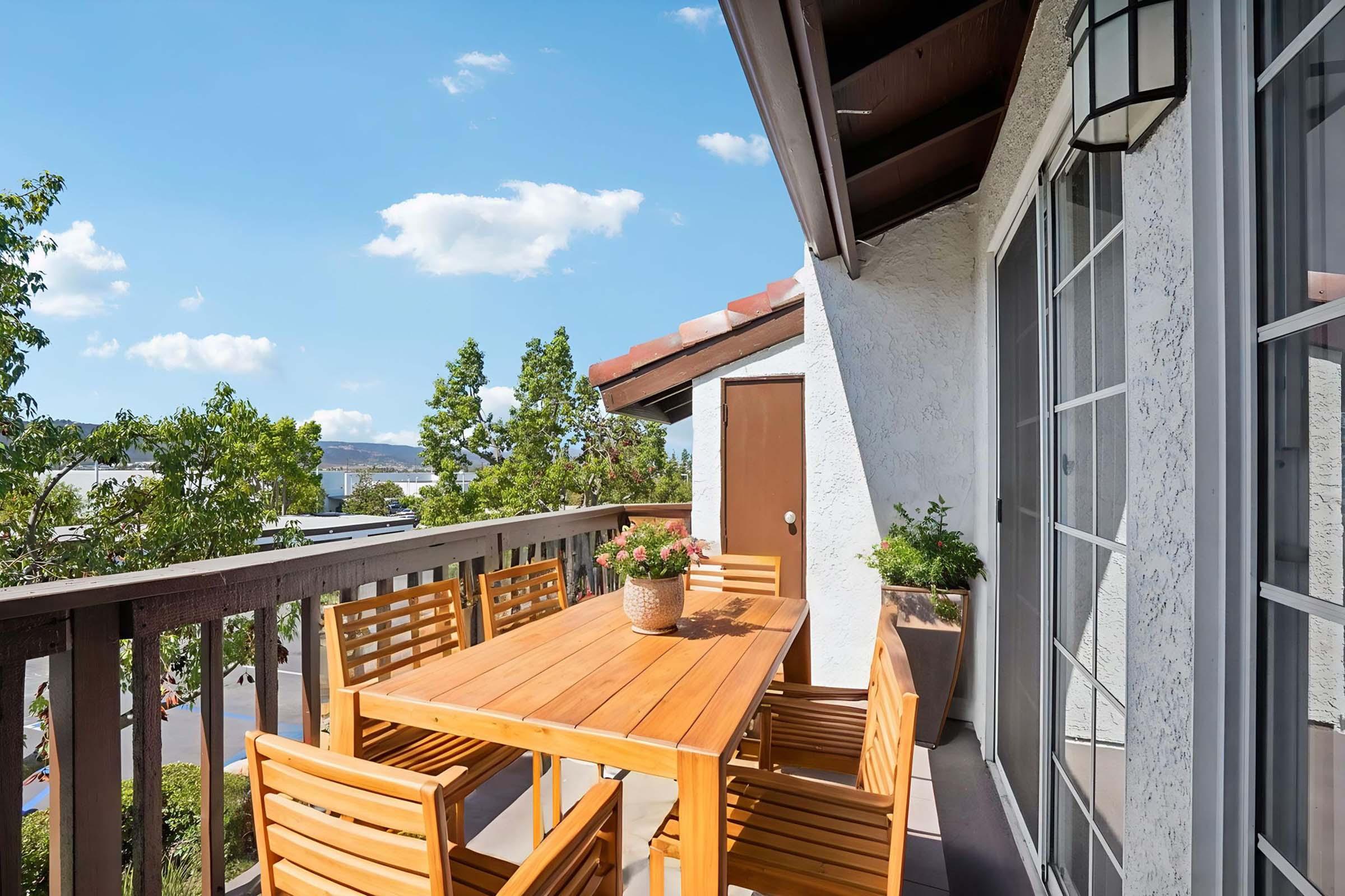 A sunny balcony with a wooden dining table surrounded by matching chairs. A potted plant sits on the table, and more greenery is present in planters. The railing offers a view of trees and a clear blue sky with a few clouds, creating a tranquil outdoor space ideal for dining or relaxing.