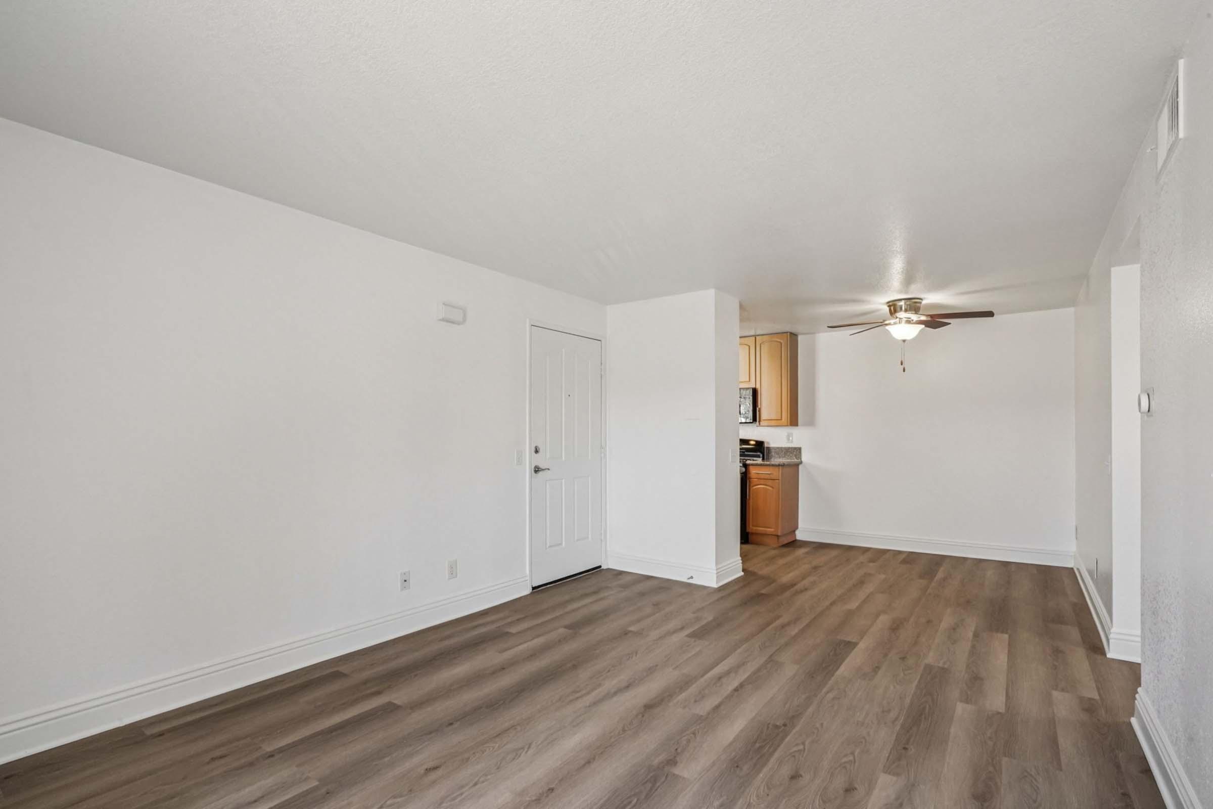 A modern, minimalistic living space with light-colored walls and wooden flooring. Features a doorway on the left, a ceiling fan, and an open kitchen area visible in the background, showcasing cabinetry and appliances. The room is well-lit and uncluttered, creating a spacious feel.