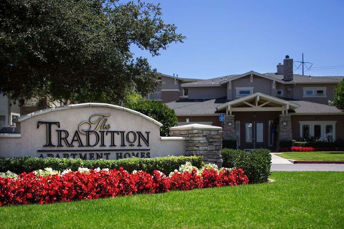 Sign for "The Tradition Apartment Homes" in front of a well-maintained apartment complex. The entrance features landscaped gardens with vibrant red flowers and green grass, set against a clear blue sky. A cozy building with a modern design is visible in the background.