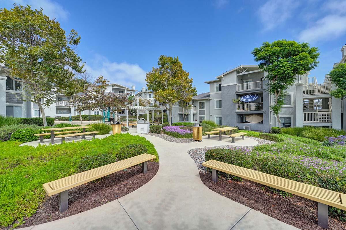 A landscaped courtyard with benches, flowering plants, and trees. Surrounding multi-story apartment buildings are visible in the background under a clear blue sky. The pathway curves through the greenery, creating a welcoming and serene atmosphere.