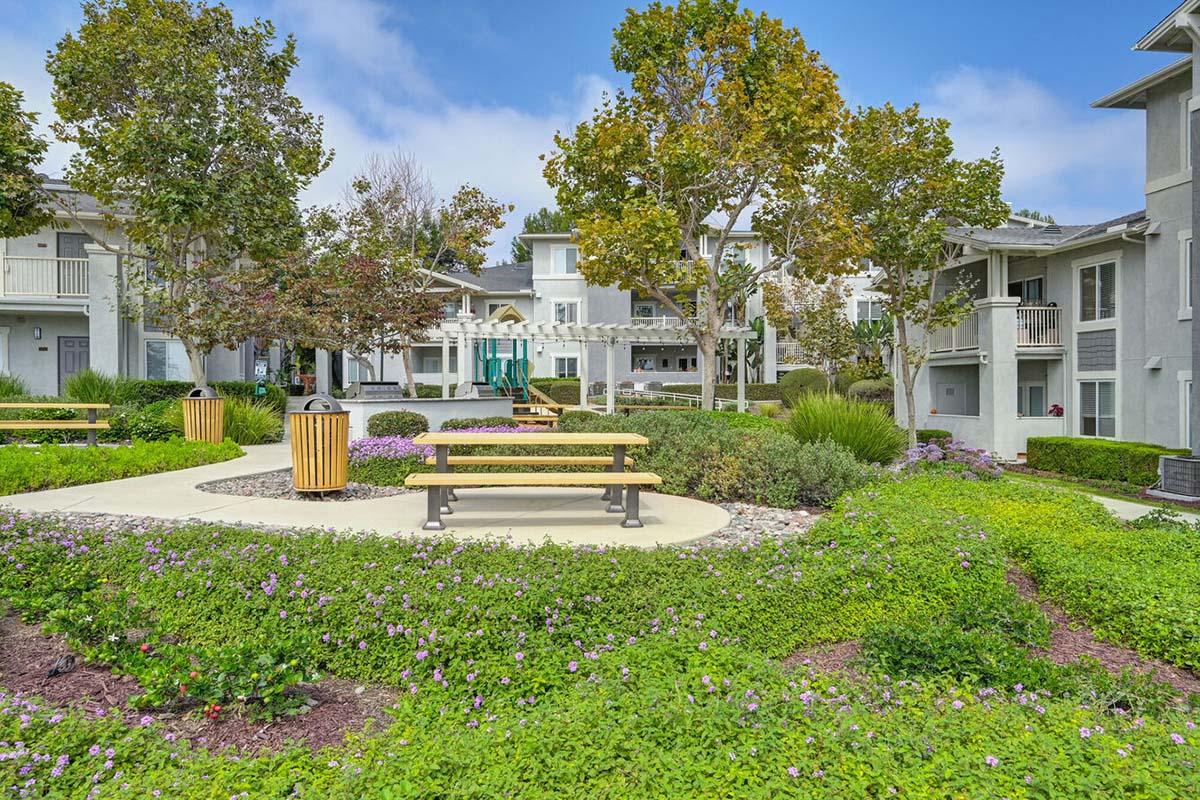 A landscaped garden area featuring benches, picnic tables, and vibrant flowers. In the background, there are residential buildings with balconies, surrounded by trees and a blue sky. Ideal for relaxation and outdoor activities.