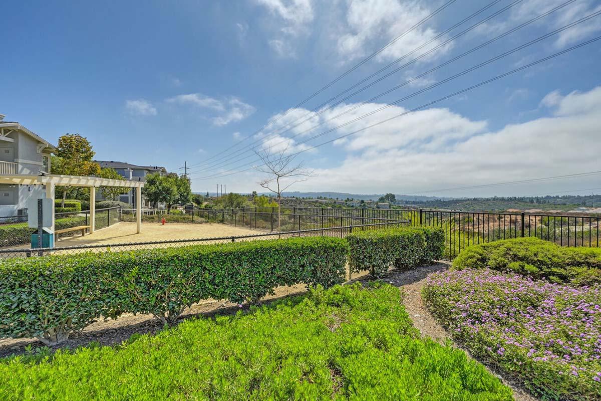 A serene landscape view showing neatly trimmed hedges and colorful flowering plants in the foreground, with a grassy area and a wooden pergola nearby. In the background, rolling hills are visible under a partly cloudy sky, along with power lines and distant trees, creating a peaceful outdoor environment.