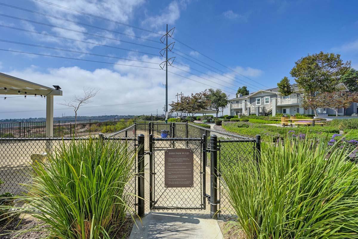 A pathway leading through a fenced area with tall grass and shrubs, leading to a sign. In the background, residential buildings and utility poles are visible under a partly cloudy sky. The scene depicts a tranquil outdoor space.