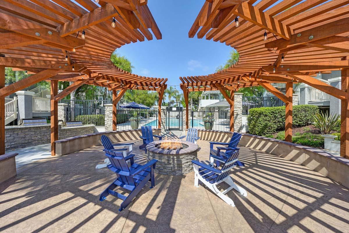 A landscaped outdoor space featuring a circular area with wooden trellises overhead, surrounded by stone seating and blue adirondack chairs. In the center, there's a fire pit. The background shows a pool area and greenery, under a clear blue sky.