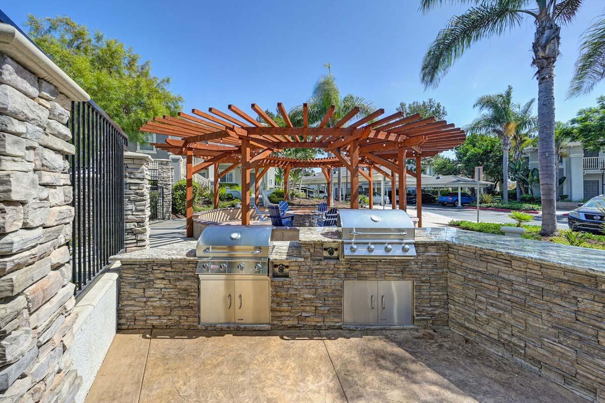 Outdoor grilling area featuring stainless steel barbecue grills under a wooden pergola. The space is surrounded by stone walls and palm trees, creating a relaxing atmosphere. Open seating is available nearby, with a view of a sunny residential area.