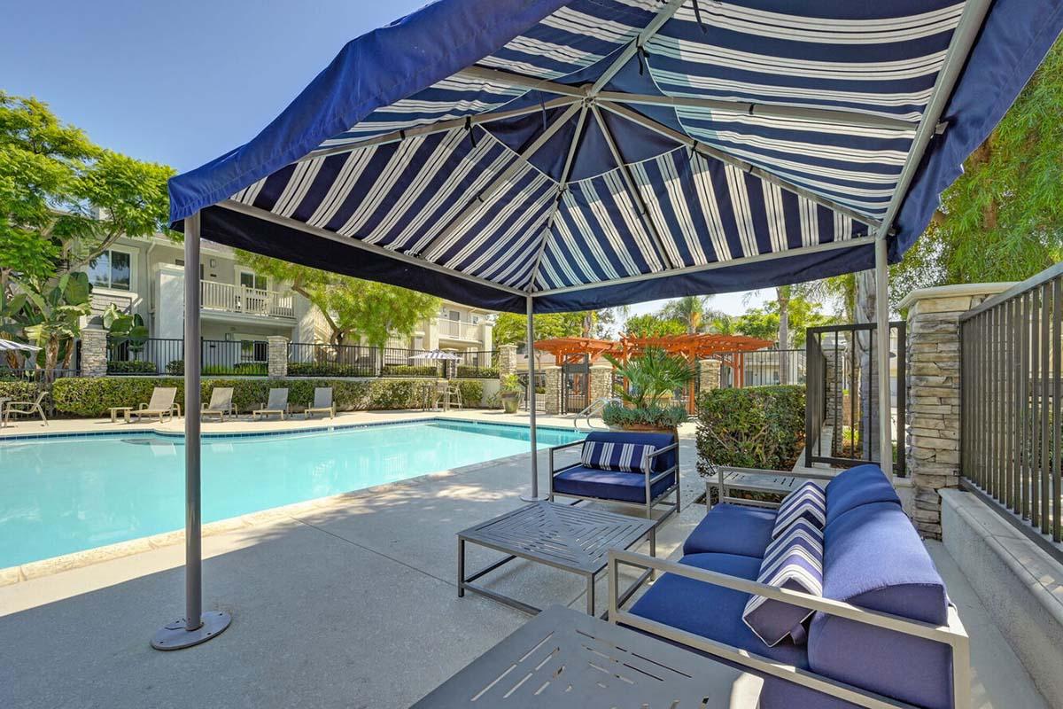 A shaded seating area beside a swimming pool, featuring a striped canopy, blue lounge chairs, and a well-maintained landscape with green trees and shrubs. In the background, there are additional lounge chairs and a fence surrounding the pool area. The scene conveys a relaxing outdoor environment.