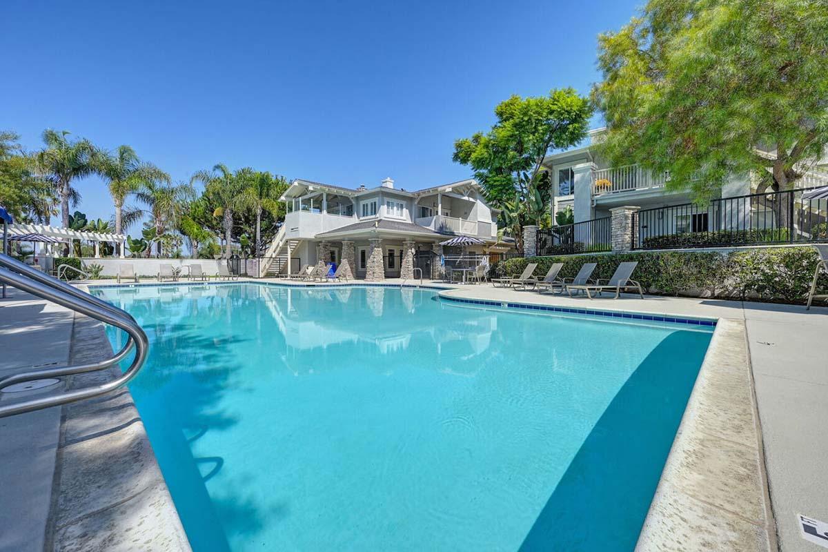 A clear blue swimming pool surrounded by lounge chairs and palm trees, with two buildings in the background and a bright blue sky overhead. The scene conveys a relaxed, sunny atmosphere ideal for leisure and enjoyment.