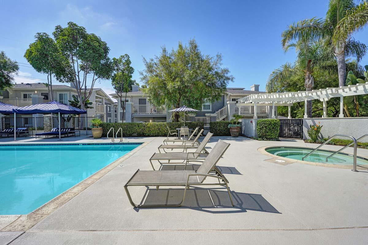 A serene pool area featuring a crystal-clear swimming pool surrounded by lounge chairs. Lush greenery and palm trees enhance the atmosphere. A hot tub is nearby, and shaded seating areas are provided with striped awnings. The scene conveys a relaxing, inviting environment perfect for leisure.