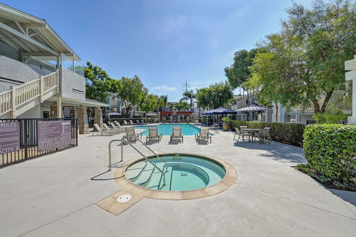 A serene outdoor pool area featuring a turquoise pool surrounded by lounge chairs, shaded by umbrellas. A hot tub is situated nearby, with landscaped greenery and a modern building in the background, creating a relaxing atmosphere for leisure and social gatherings.