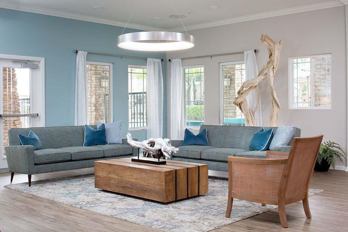 A stylish living room featuring a blue accent wall, a modern circular light fixture, a gray sofa with decorative blue pillows, and a wooden coffee table. There is also a single armchair, a decorative piece on the table, and large windows allowing natural light to fill the space.