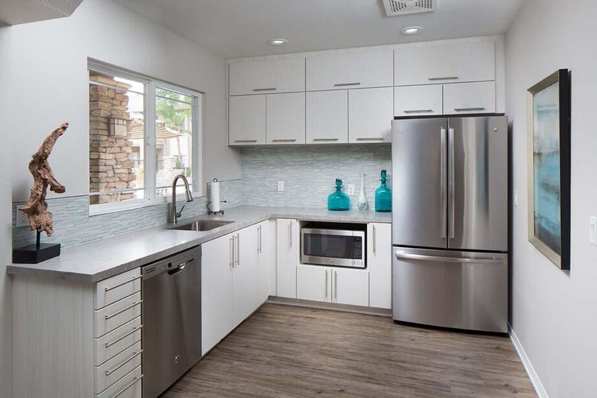 Modern kitchen featuring sleek white cabinetry, stainless steel appliances including a refrigerator and microwave, and a light gray countertop. Natural light streams through a window, illuminating decorative teal vases and a sculptural wooden accent piece on the countertop. Flooring is a warm wood tone.