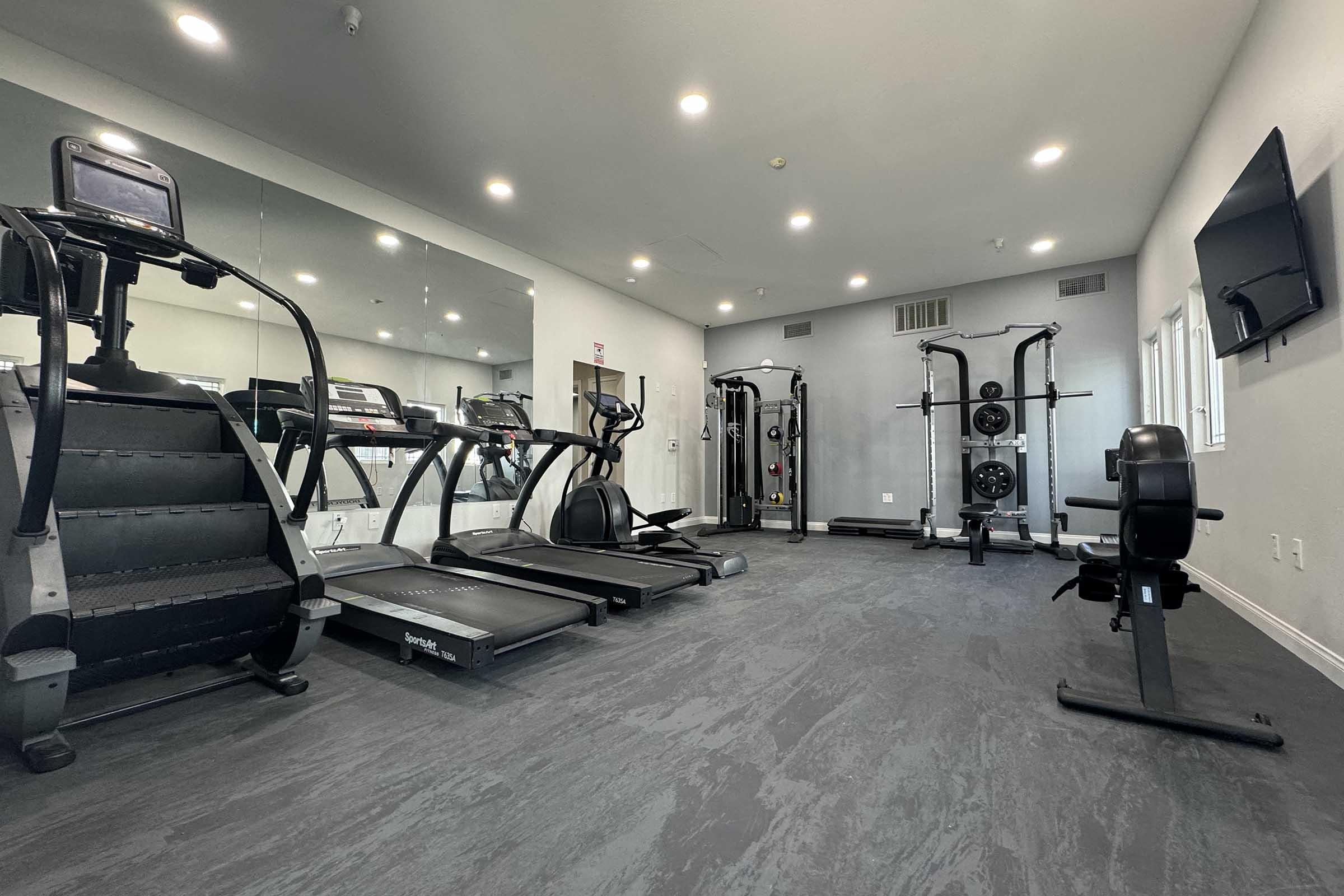 A modern gym interior featuring various exercise equipment including treadmills, a stationary bike, a strength training machine, and a weight bench. The space is well-lit with overhead lights and mirrors, creating an inviting atmosphere for workouts. A television is mounted on the wall for entertainment.