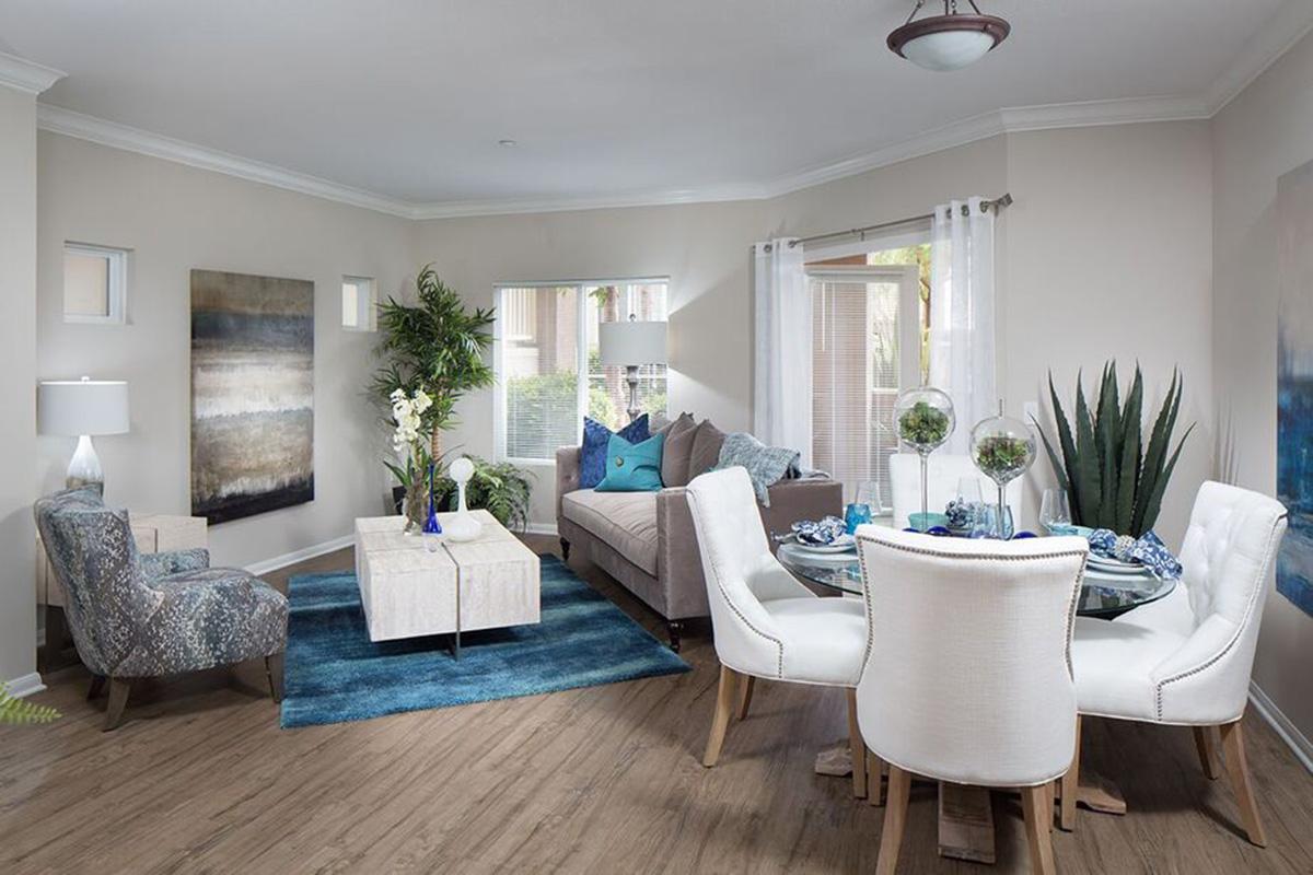 A modern living room featuring a light color scheme with beige walls and hardwood flooring. The space includes a comfortable gray sofa, a round dining table with white chairs, a decorative coffee table, and various plants. Decorative accents in blue and white enhance the tranquil atmosphere.