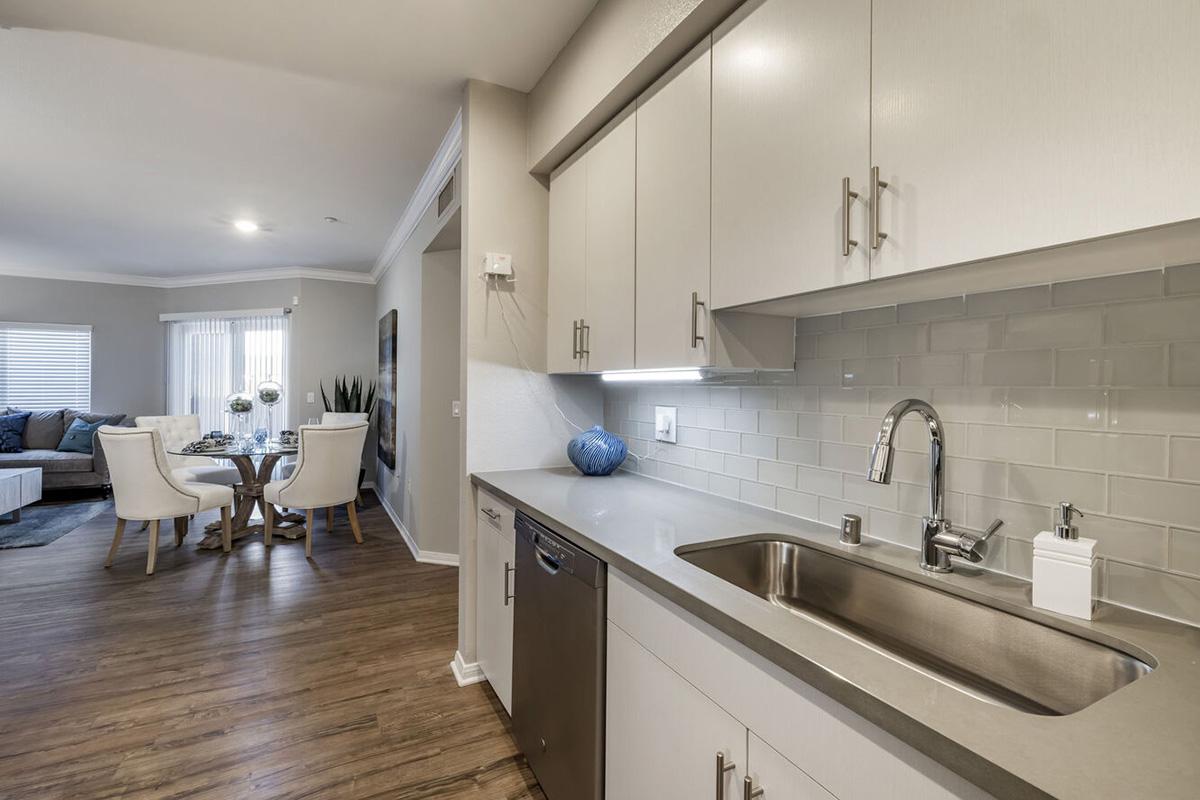 Modern kitchen with sleek cabinetry, stainless steel sink, and appliances. Light-colored countertops complement stylish backsplash. A dining area is visible in the background, featuring a round table and white chairs, with a large window allowing natural light to illuminate the space.