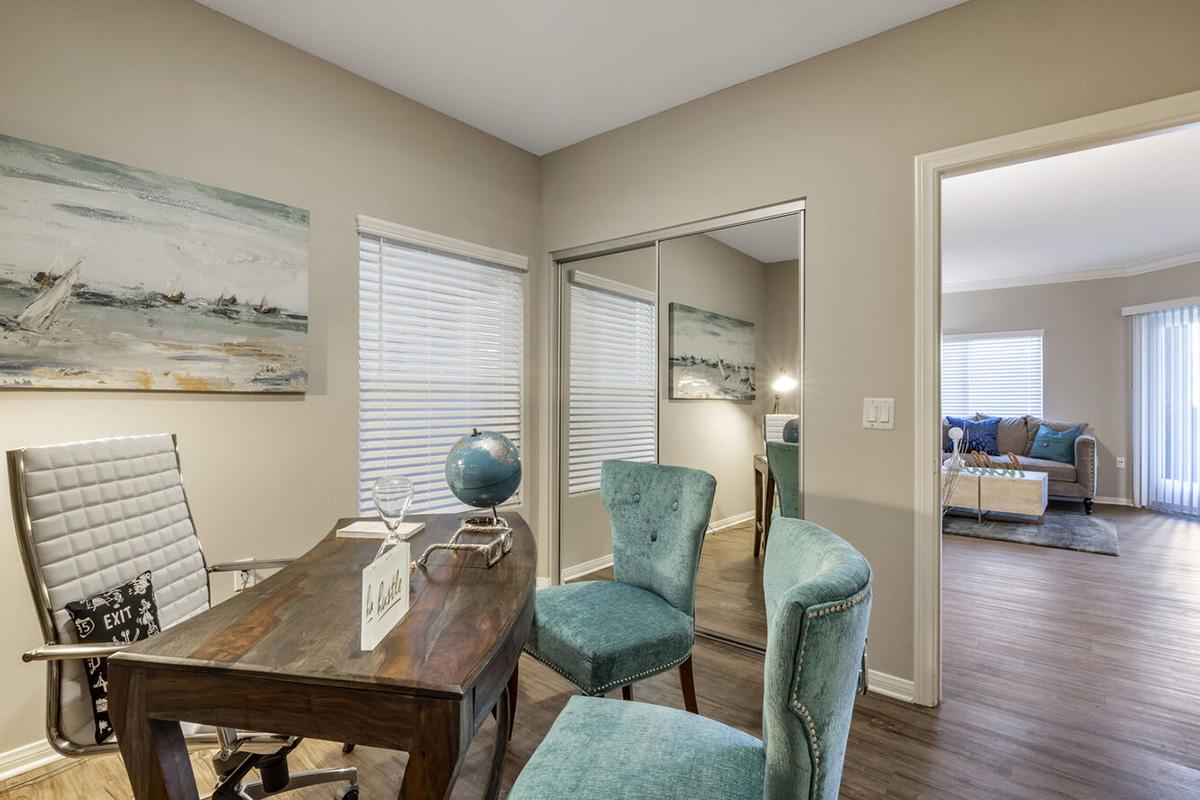 A stylish home office featuring a wooden desk with two turquoise chairs, a globe, and decorative items. There's a large mirror reflecting the room, and large windows with blinds let in natural light. In the background, a cozy living area with a sofa and decorative cushions is visible.